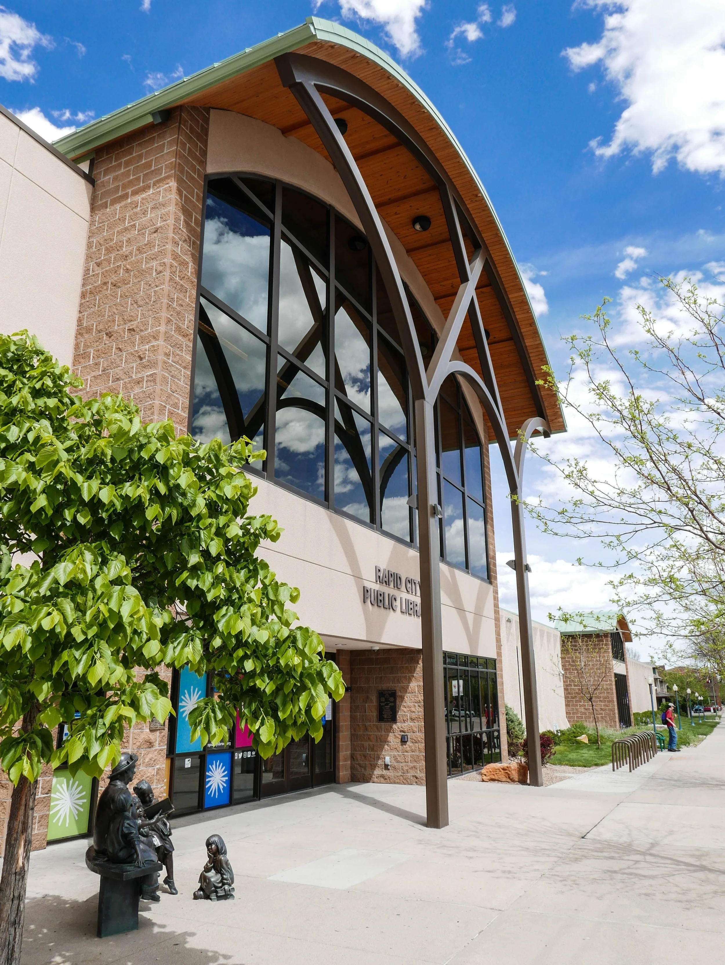 Exterior view of Rapid City Public Library with modern architectural design, large glass windows, and landscaped outdoor area with small trees and sculptures.