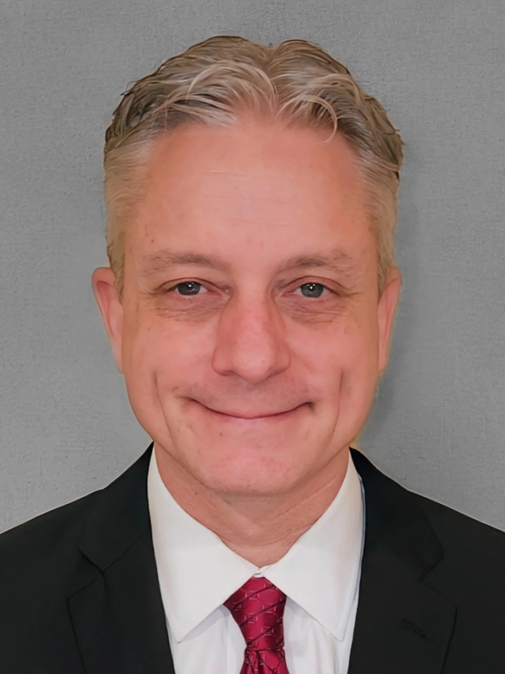 A professional headshot of a middle-aged man with blonde hair wearing a black suit, white shirt, and red tie, smiling in front of a gray background.