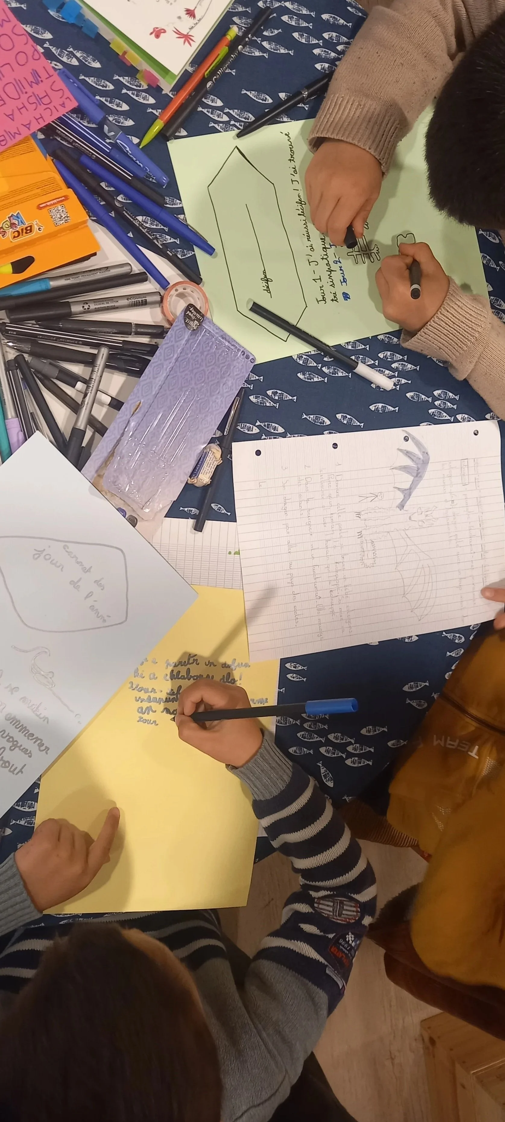 Atelier d'écriture enfants sur le thème des sardinosaures. Des feuilles de papier colorées avec différents stylos, des crayons, des notes autocollantes pour stimuler la créativité.