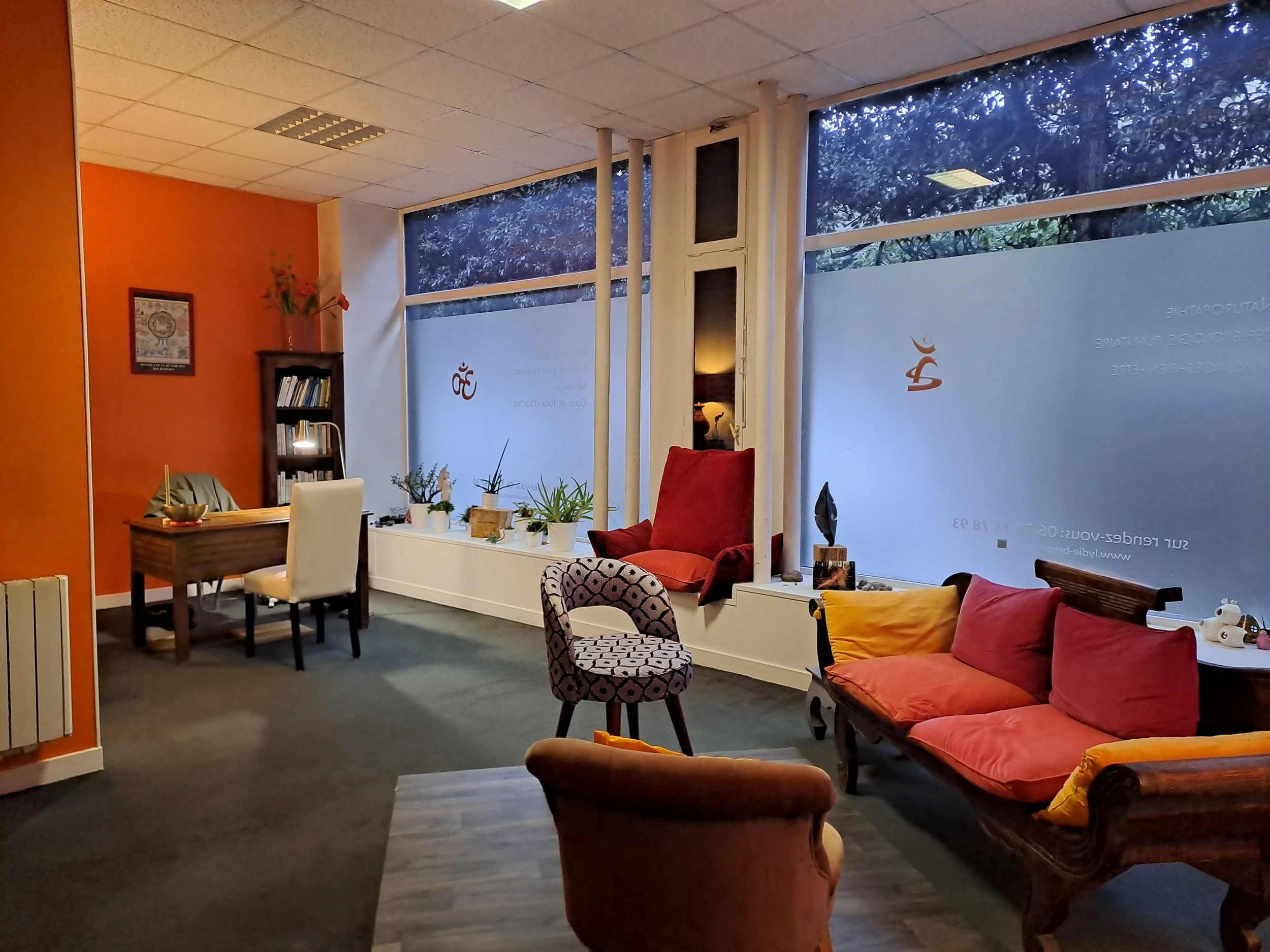 Salon chaleureux avec canapé rouge, fauteuils, table et plantes, grands fenêtres laissant entrer la lumière naturelle.