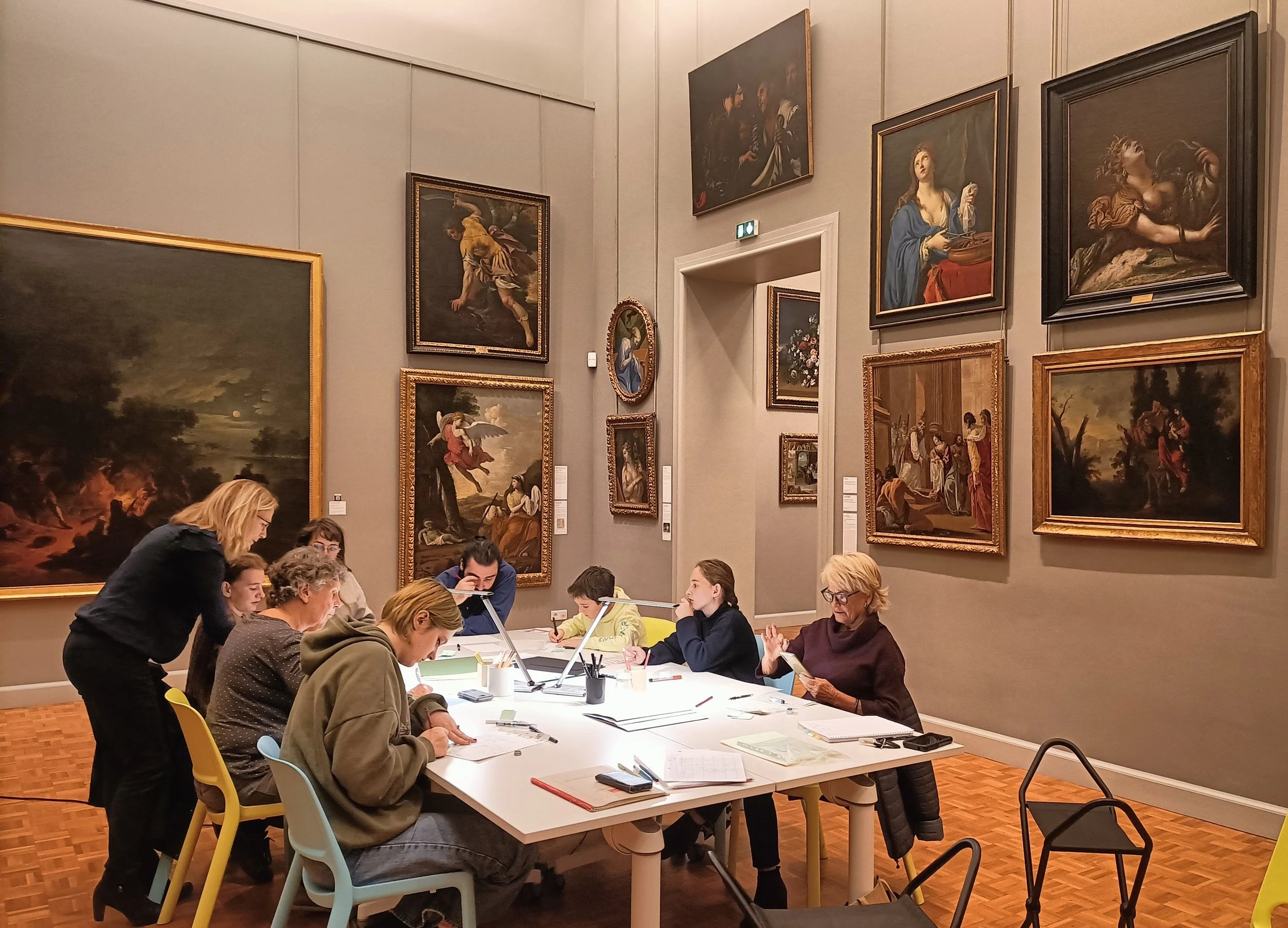 Groupe d'atelier d'écriture au musée, des personnes assises autour d'une table dans un musée, observant et écrivant autour des œuvres d'art suspendues aux murs.