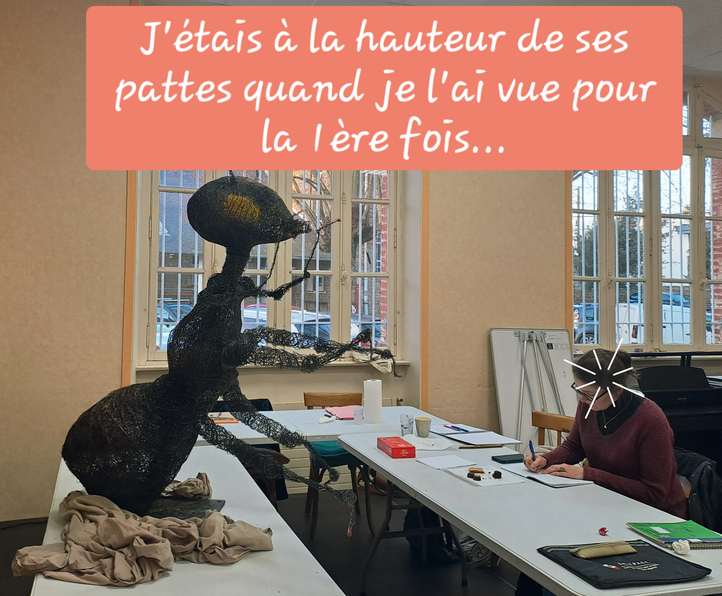Une sculpture en fil métallique représentant une fourmi géante face à des participants de l'atelier d'écriture assis à une table. La scène est dans une pièce avec des grandes fenêtres et une décoration simple.
