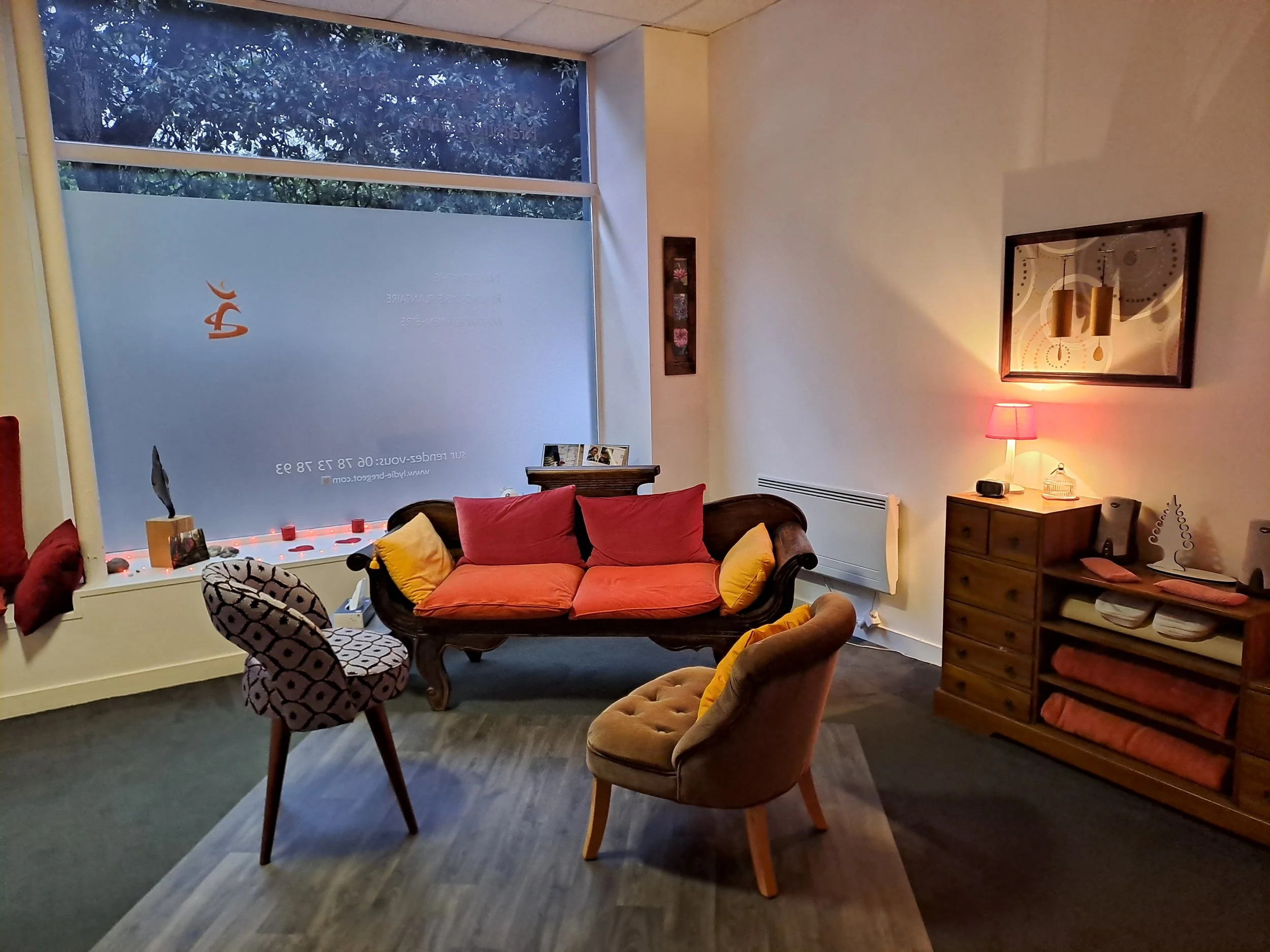 Salon d'intérieur avec canapé en bois et coussins roses et jaunes, deux fauteuils en tissu beige et motifs, lampe avec abat-jour rose, meuble en bois avec piles de linge et haut-parleurs, mur décoré avec tableau