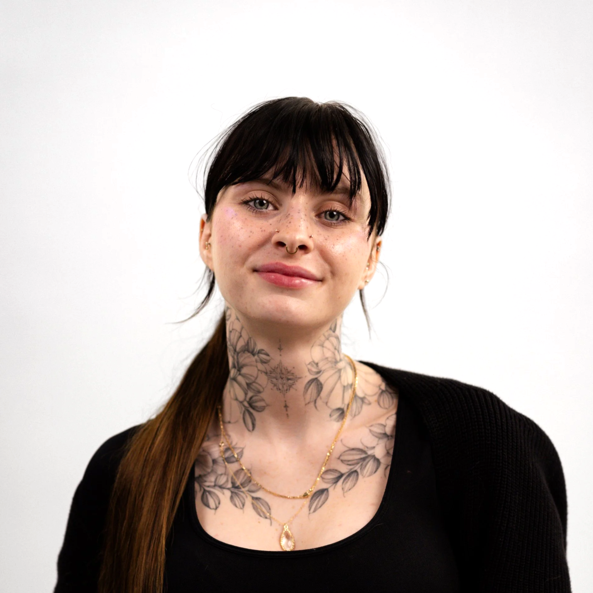 Portrait of a smiling woman with black hair, tattoos on neck and chest, wearing a black top and gold jewelry against a plain white background.