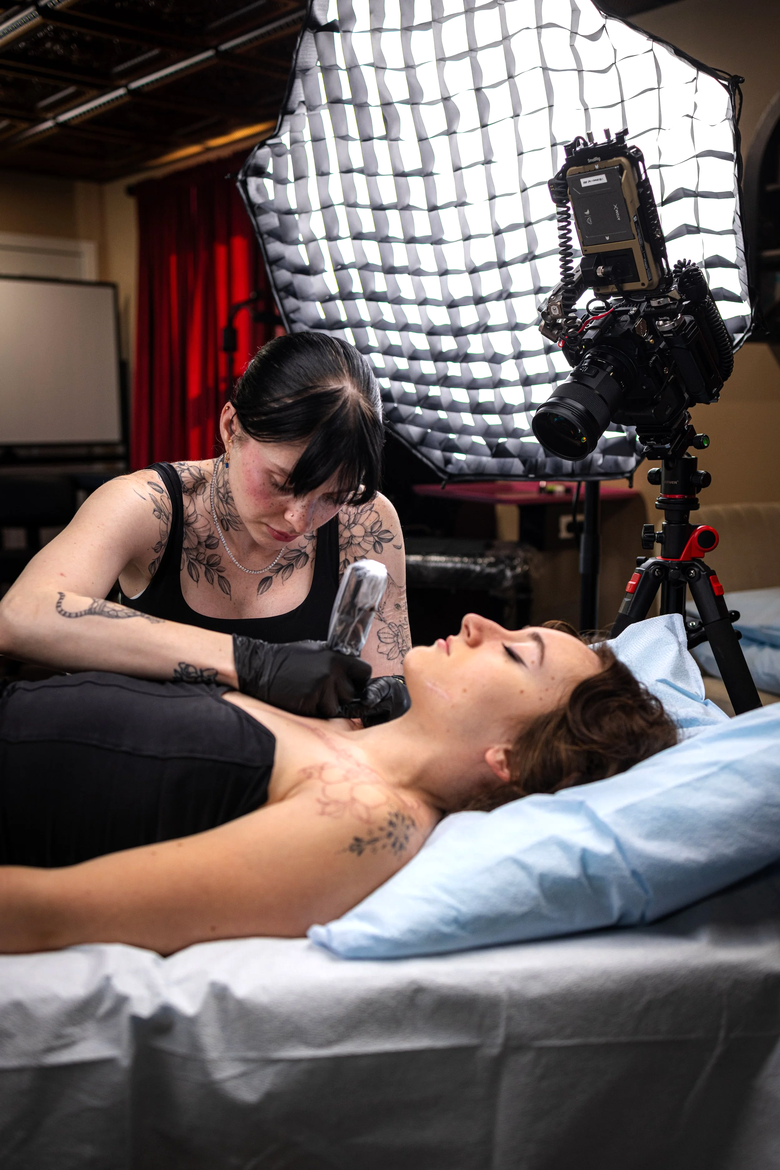 A tattooed woman lying on a hospital bed while a tattoo artist creates a tattoo on her chest, with professional lighting and camera setup involved in the process.