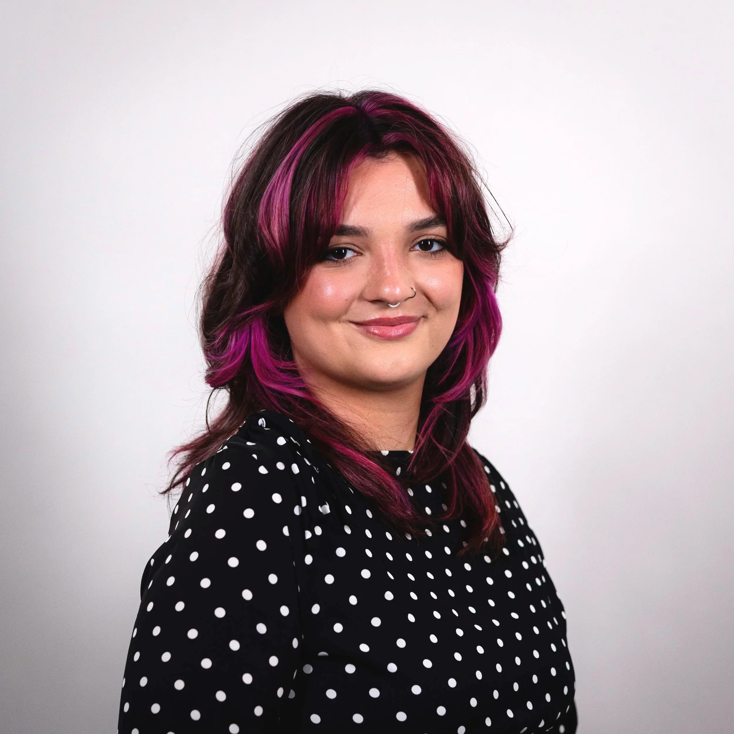 A young woman with purple hair and a black polka dot shirt smiling against a plain white background.