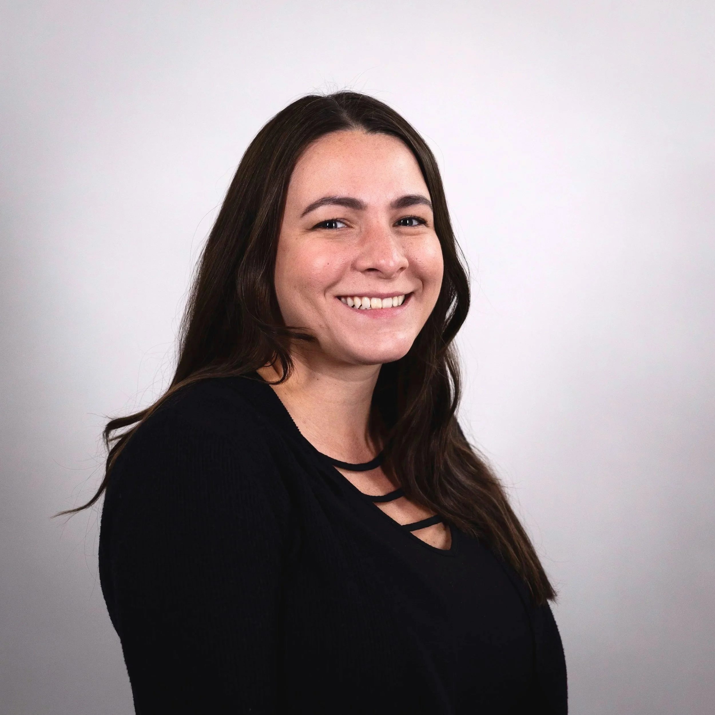 Portrait of a smiling woman with long brown hair, wearing a black top, against a plain gray background.