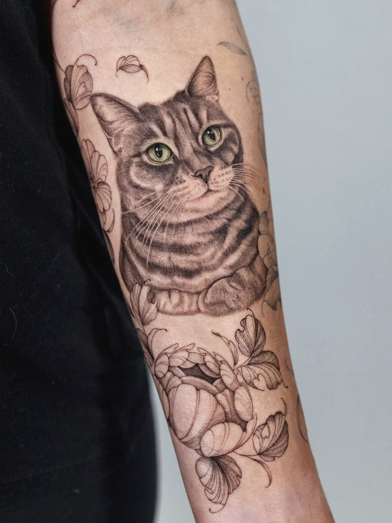 A detailed black and gray tattoo of a cat with green eyes, surrounded by flowers and leaves, on a person's arm.