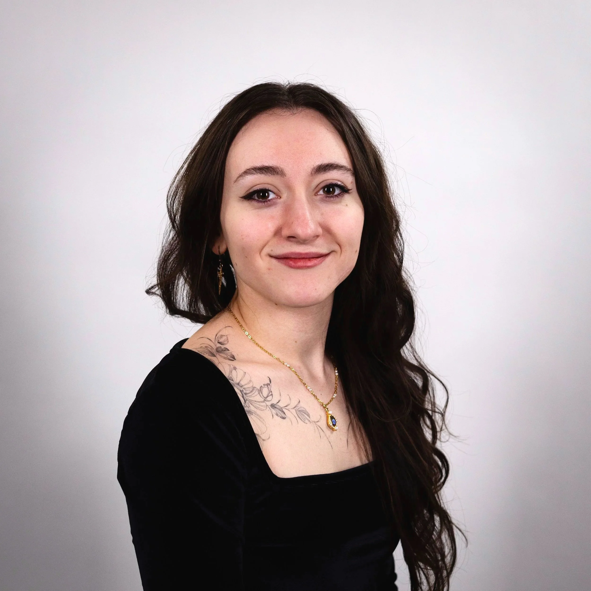 Portrait of a young woman with long wavy dark hair, wearing a black top, gold necklace with blue gemstone, and earrings, standing against a plain white background.