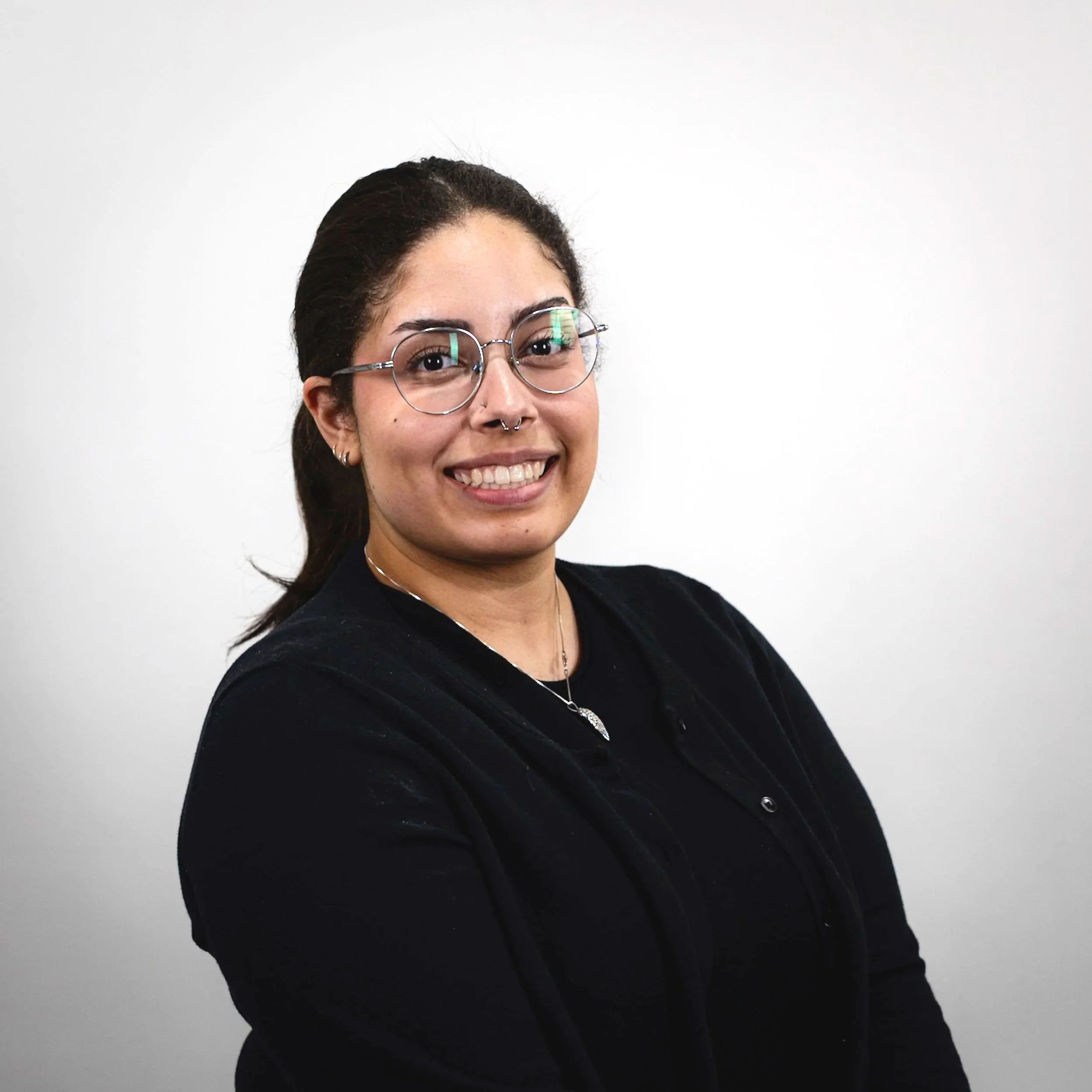 A young woman with glasses and a septum piercing smiling, wearing a black top and jacket, against a plain white background.
