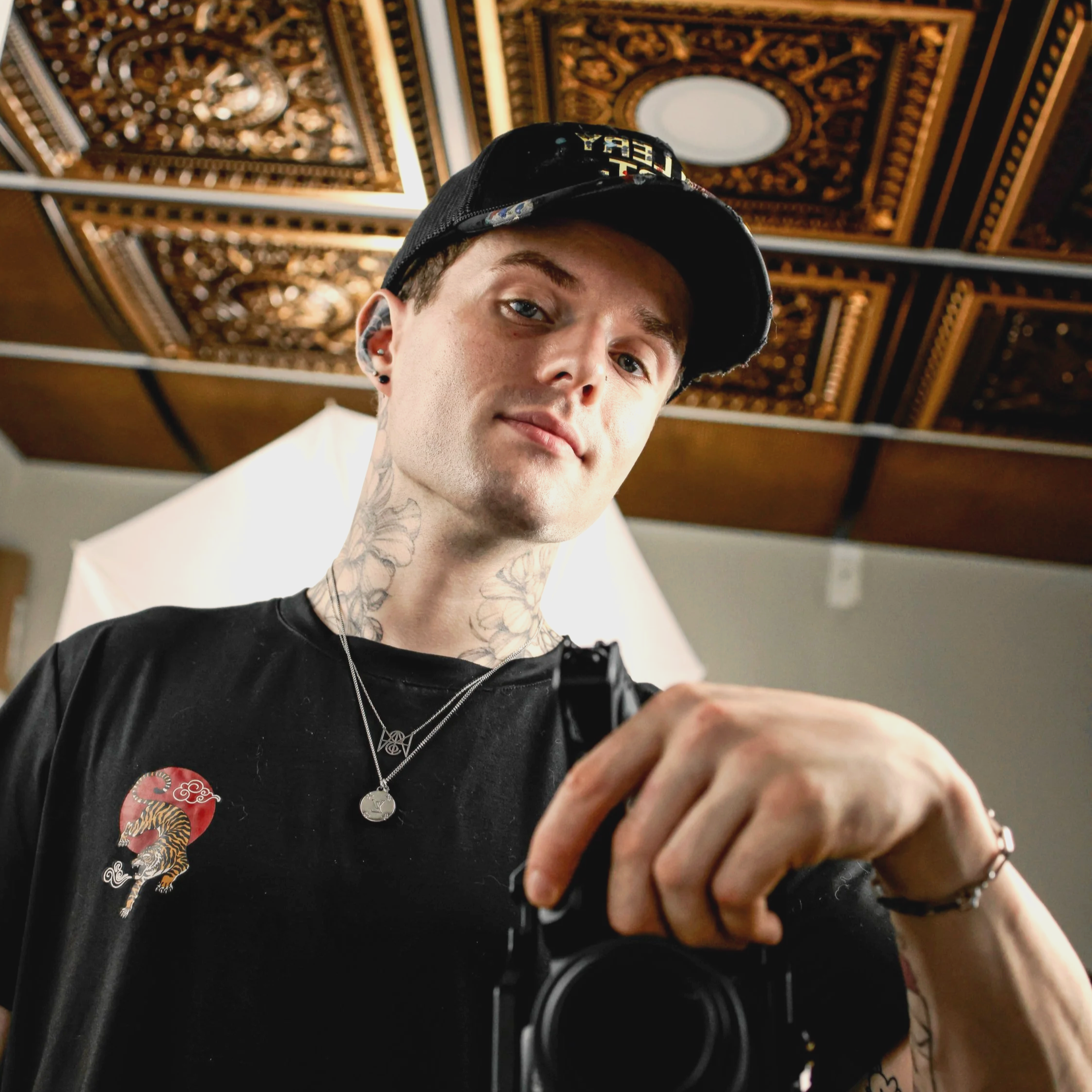 Young man with tattoos on his neck wearing a black cap and a black T-shirt, taking a selfie in a room with ornate ceiling decor and a photography backdrop.