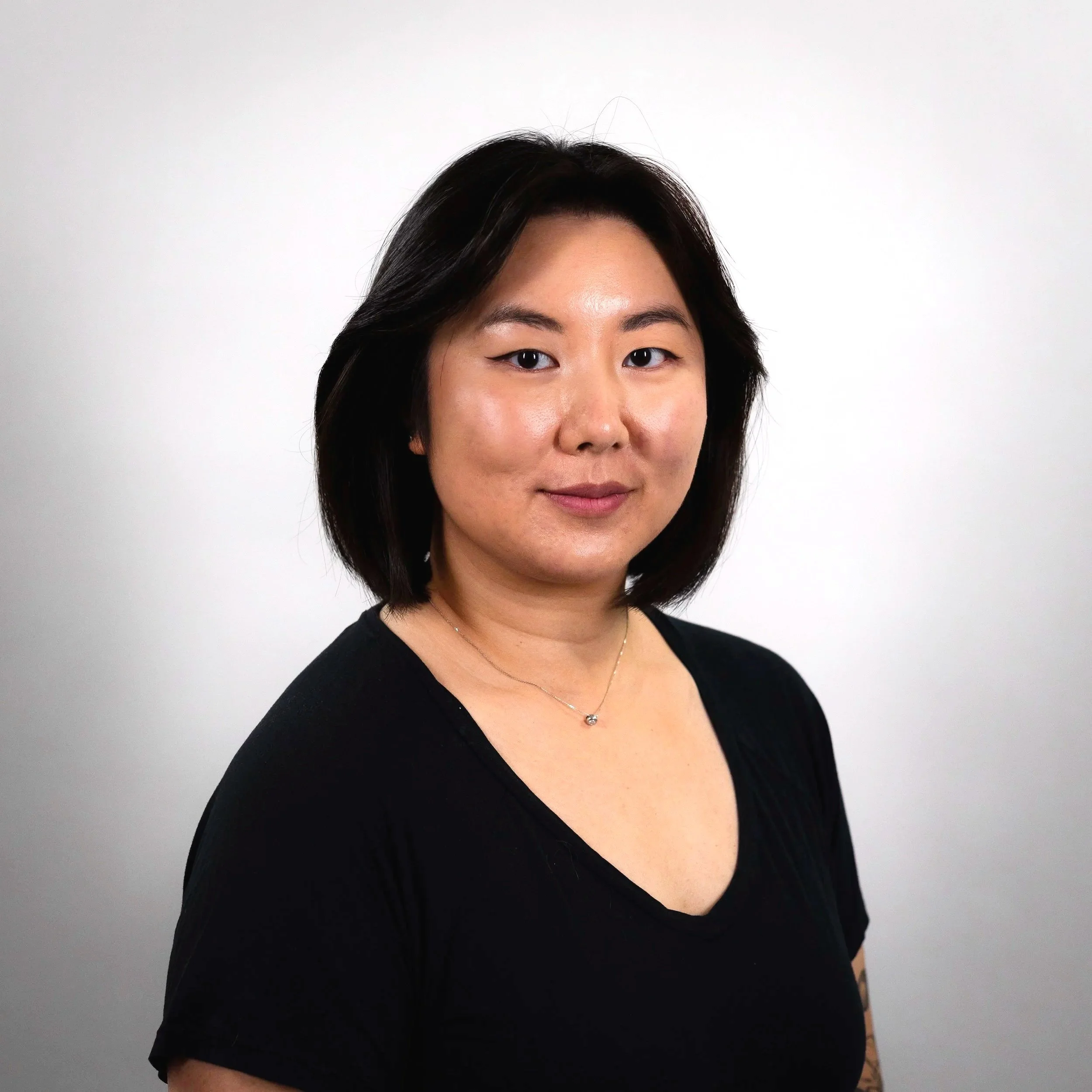 A woman with shoulder-length black hair wearing a black top and a delicate necklace, posed against a plain white background.