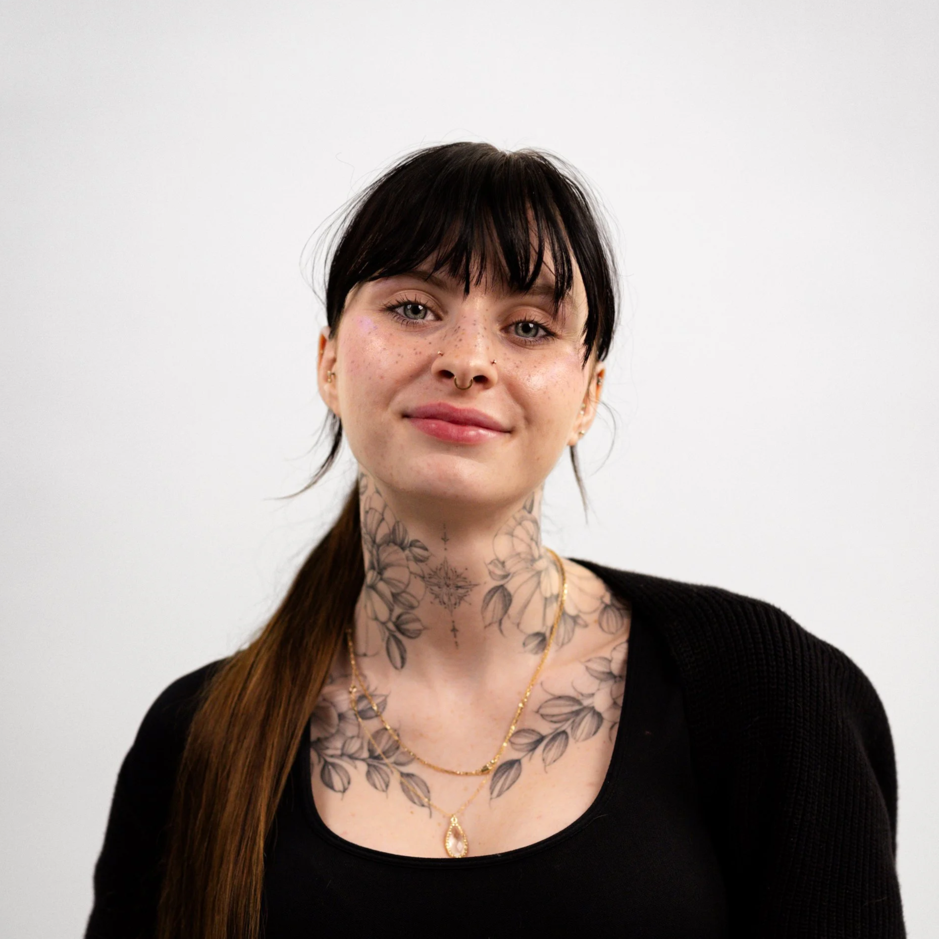 A woman with black hair, tattoos on her neck and chest, wearing a black top, gold necklaces, and earrings, standing against a plain white background.