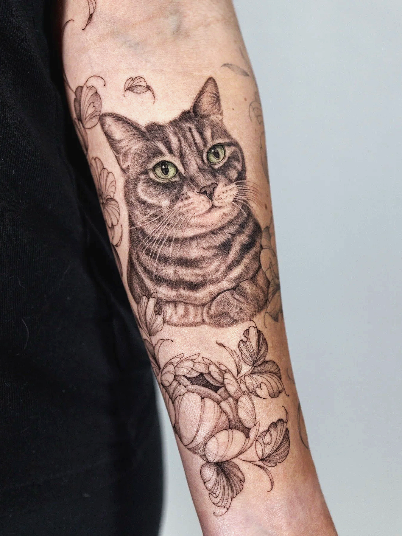 Black and gray fine line tattoo of a cat with green eyes surrounded by leaves and peonies on a person's arm.