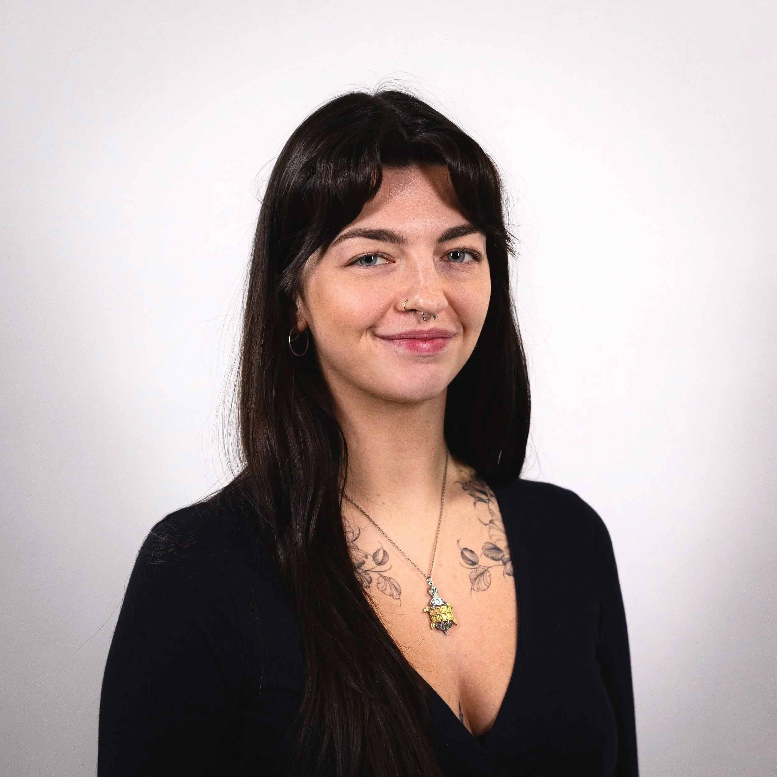 Portrait of a young woman with long dark hair, blue eyes, and light skin, wearing a black top, necklaces, earrings, and a nose ring, against a plain white background.