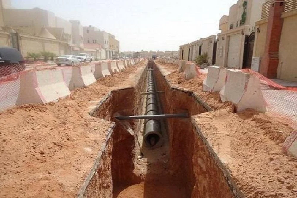 Construction site with a deep trench in a city street, with a black pipe laid at the bottom and protective barriers on either side.