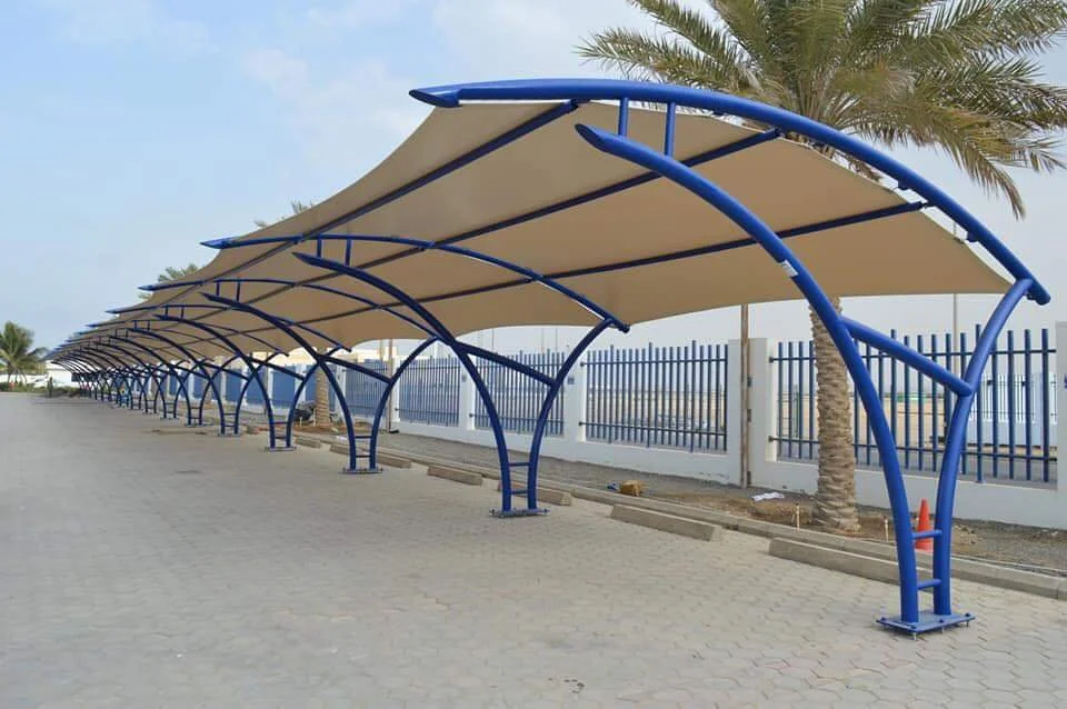 Row of modern shade structures with blue metal frames and beige fabric canopies, situated along a paved walkway behind a white and blue metal fence, with palm trees in the background.