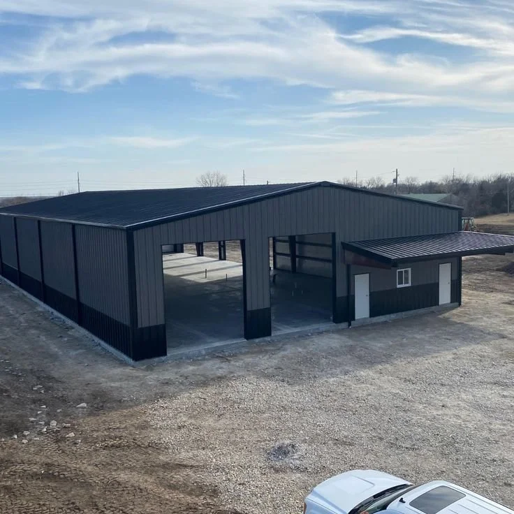 Large gray metal building, possibly a storage shed or warehouse, with a small extension on one side, situated on a gravel lot under a partly cloudy sky.