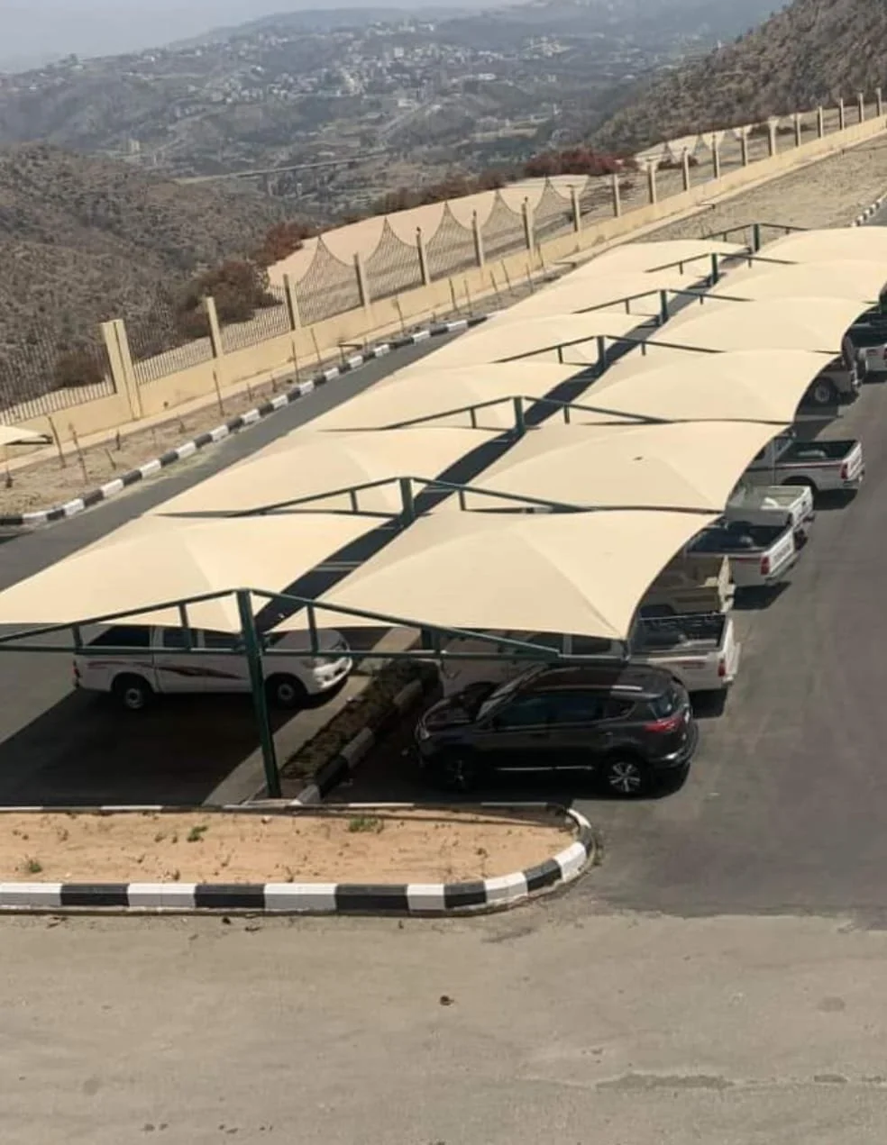 Parking lot with multiple cars parked under large beige sun shades, fencing along a hillside overlooking a landscape with mountains and valleys in the background.