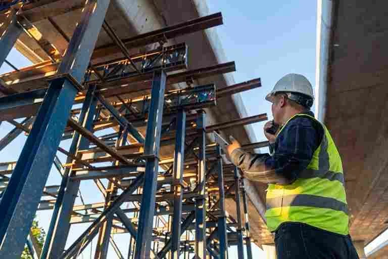 Construction worker in safety gear inspecting a steel support structure under a bridge.