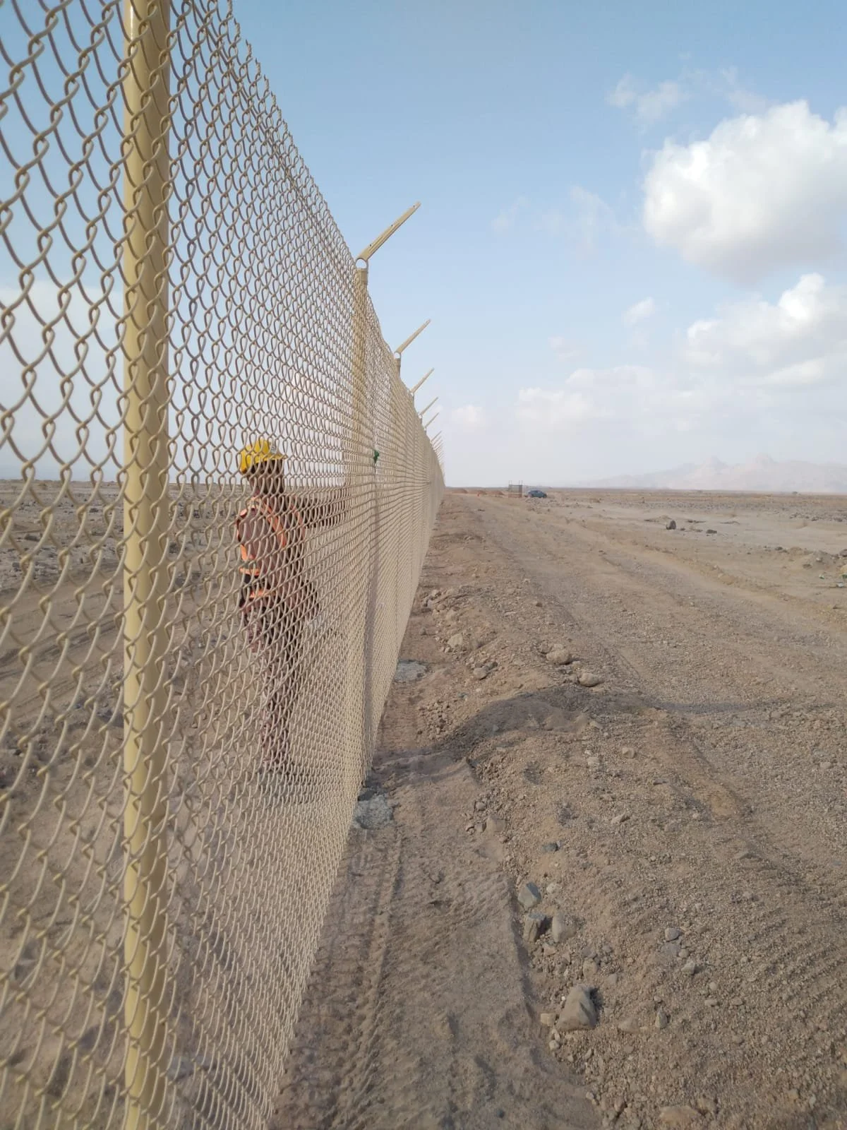 A chain-link fence with barbed wire on top stretches into the distance across a barren desert landscape. There are tire tracks in the dirt and a person wearing a helmet and sunglasses standing near the fence, with cloudy sky overhead.