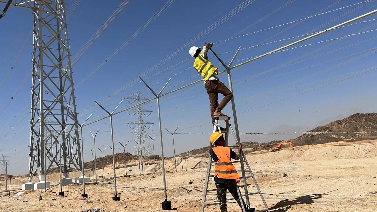 Two workers working on power lines and infrastructure in a desert-like area with wind turbines in the background.