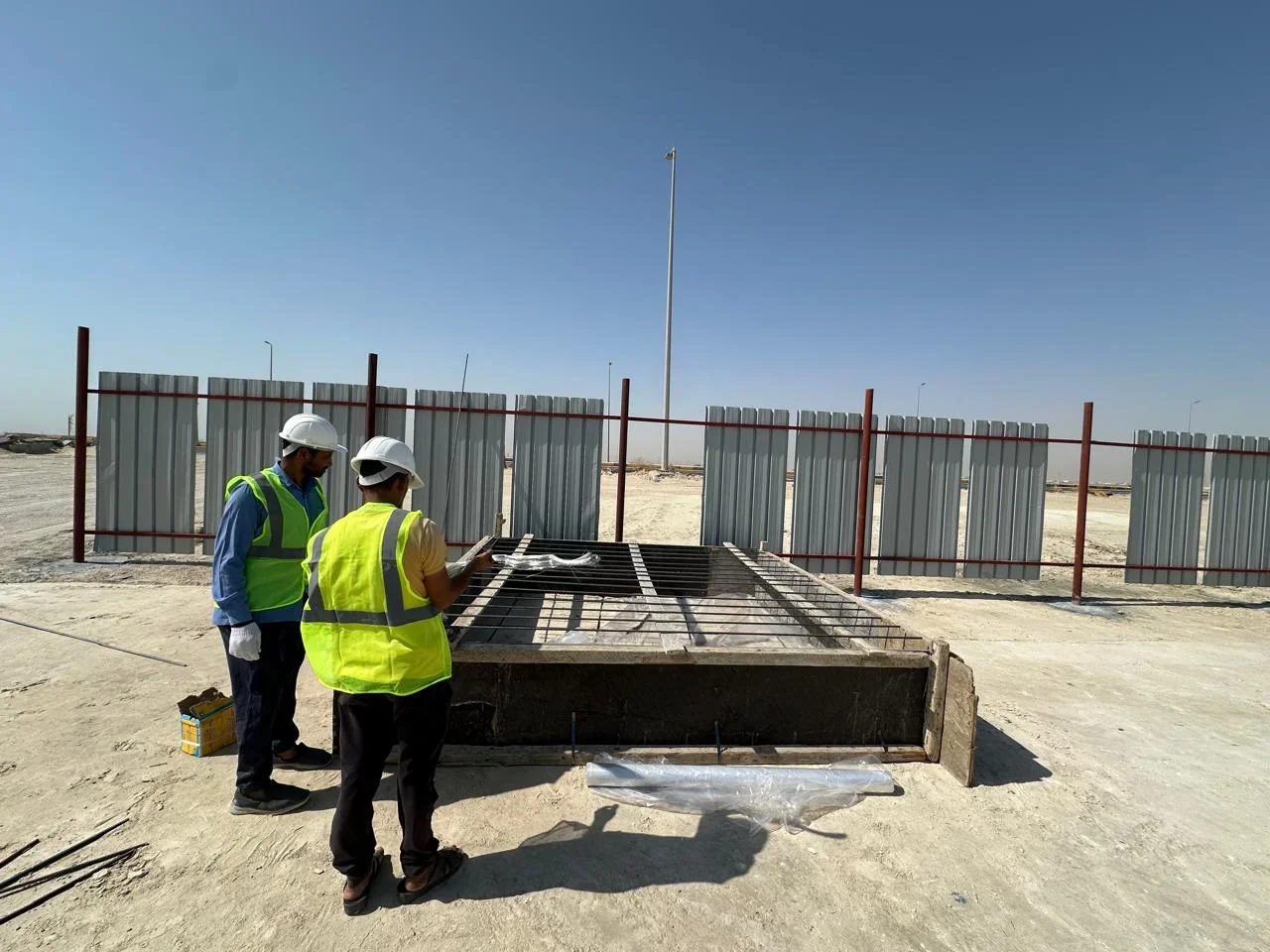 Two construction workers wearing safety vests and helmets inspecting steel reinforcement on a construction site with a metal fence and clear blue sky in the background.