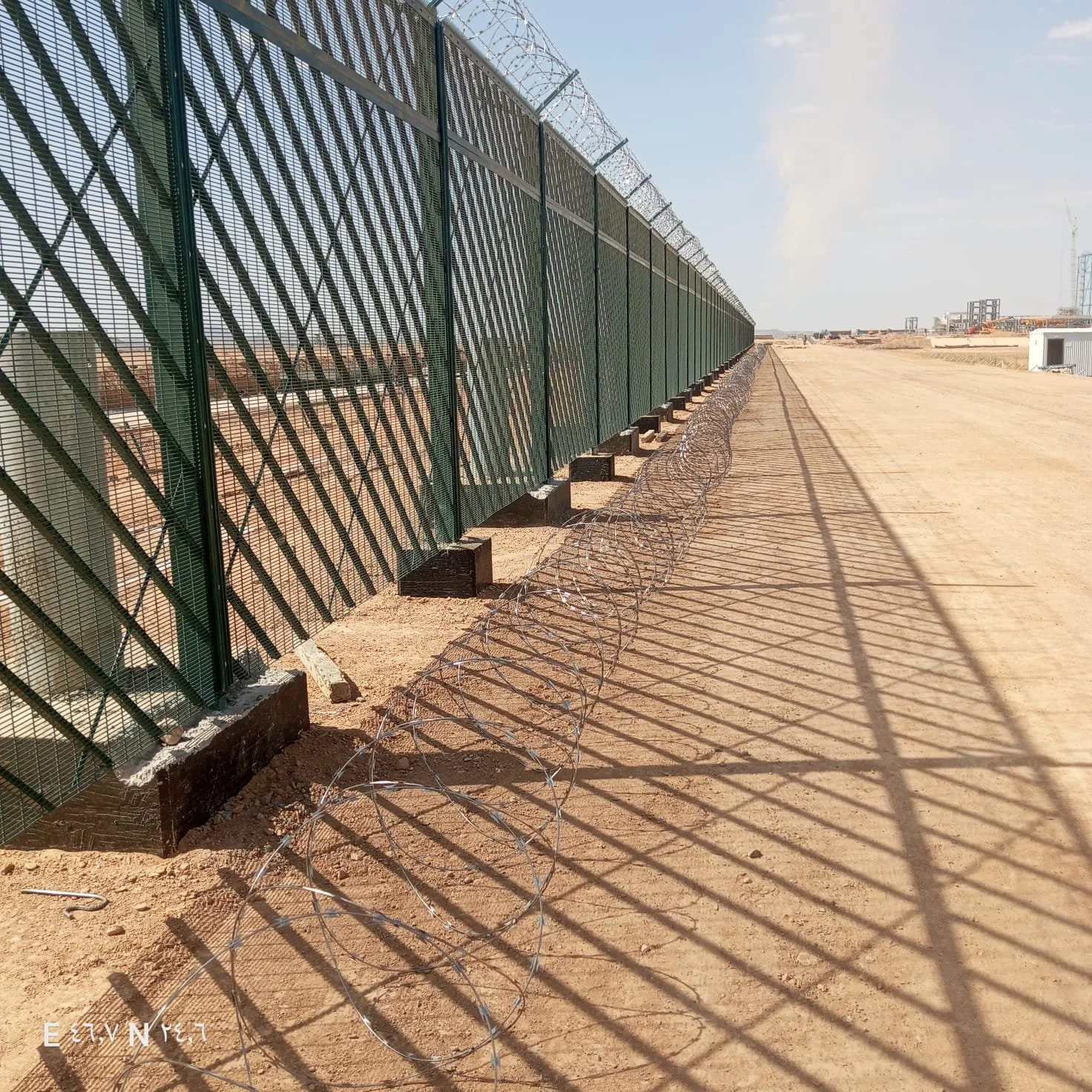 A long metal fence with barbed wire on top, casting shadows on the sandy ground nearby.