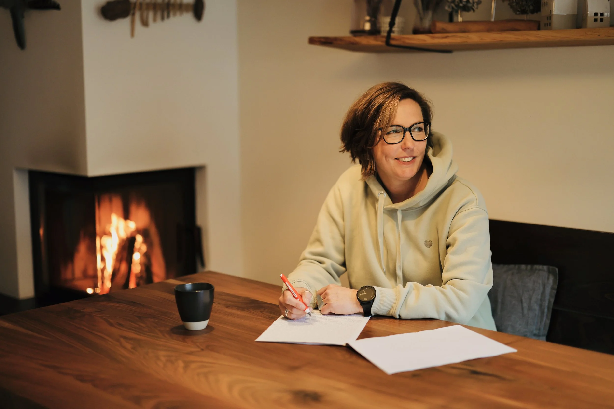 Julia Fieweger sitzt am Tisch neben einem Kaminfeuer und korrigiert handschriftlich ein Manuskript aus dem Bereich Reiseliteratur oder Biografien