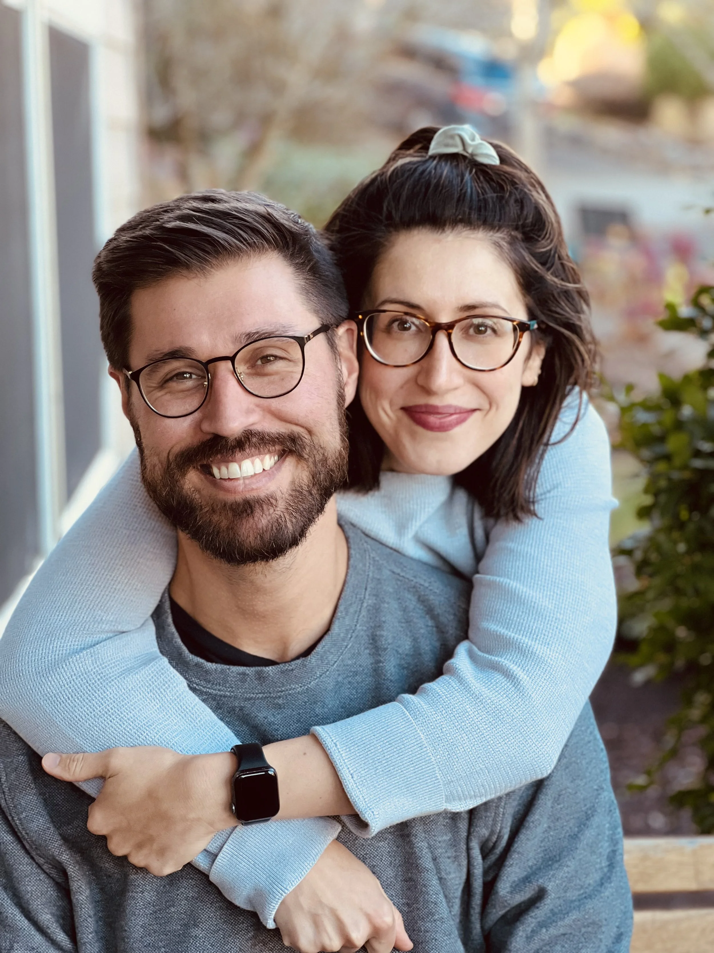 A smiling man with glasses and a beard is giving a piggyback ride to a woman with glasses, dark hair, and a hair tie, outside near a window with a blurred autumn background.