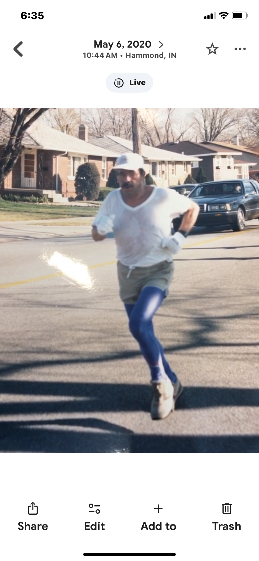 A man running on a residential street, dressed in athletic gear including a white shirt, gray shorts, blue leggings, white hat, and sneakers, with houses and cars in the background.