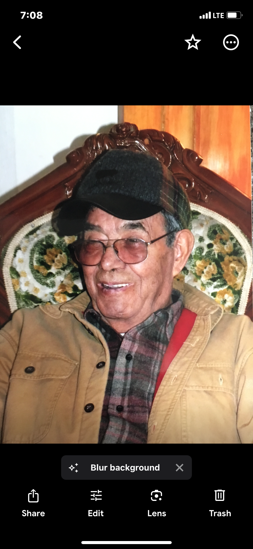 An elderly man with glasses, wearing a plaid cap and a beige jacket, sitting on a floral-patterned chair in front of a wooden panel background.