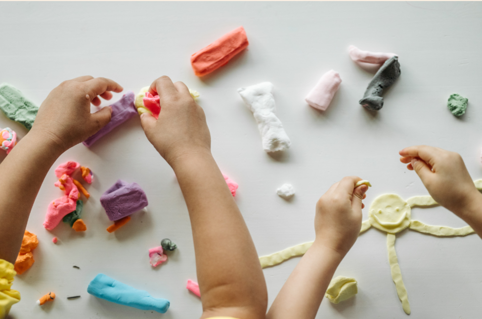 Two children playing with colorful playdough on a white surface, creating shapes and figures.