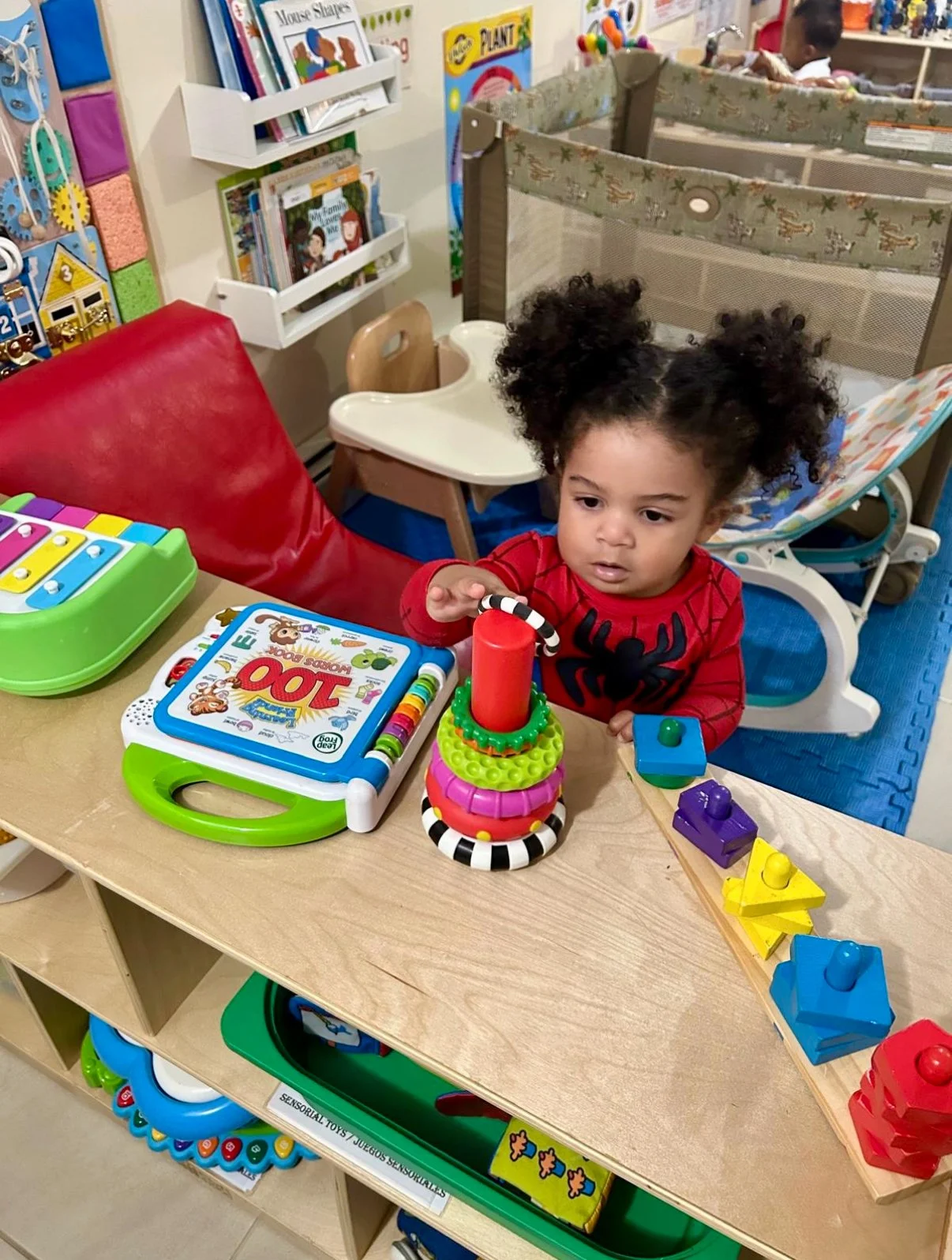 A young girl with curly hair in puffs, wearing a red shirt with a black spider graphic, playing with colorful stacking toys and blocks at a table in a playroom or classroom.