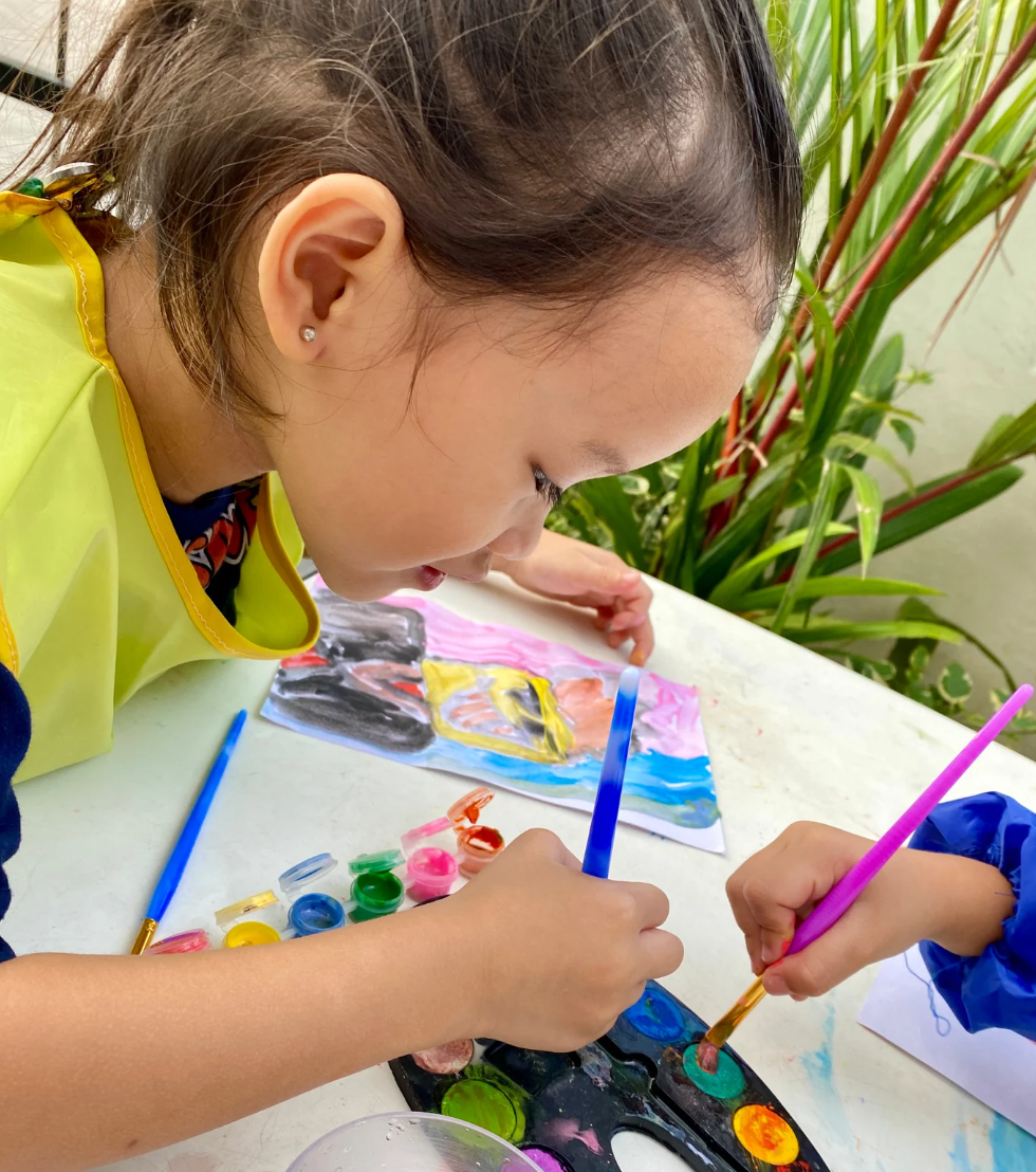 A young girl with earrings, wearing a yellow apron, painting with a blue brush on a paper with colorful abstract art. Another child's hand with a pink brush is also visible, painting on a different paper. Art supplies are on the table, and there are plants in the background.