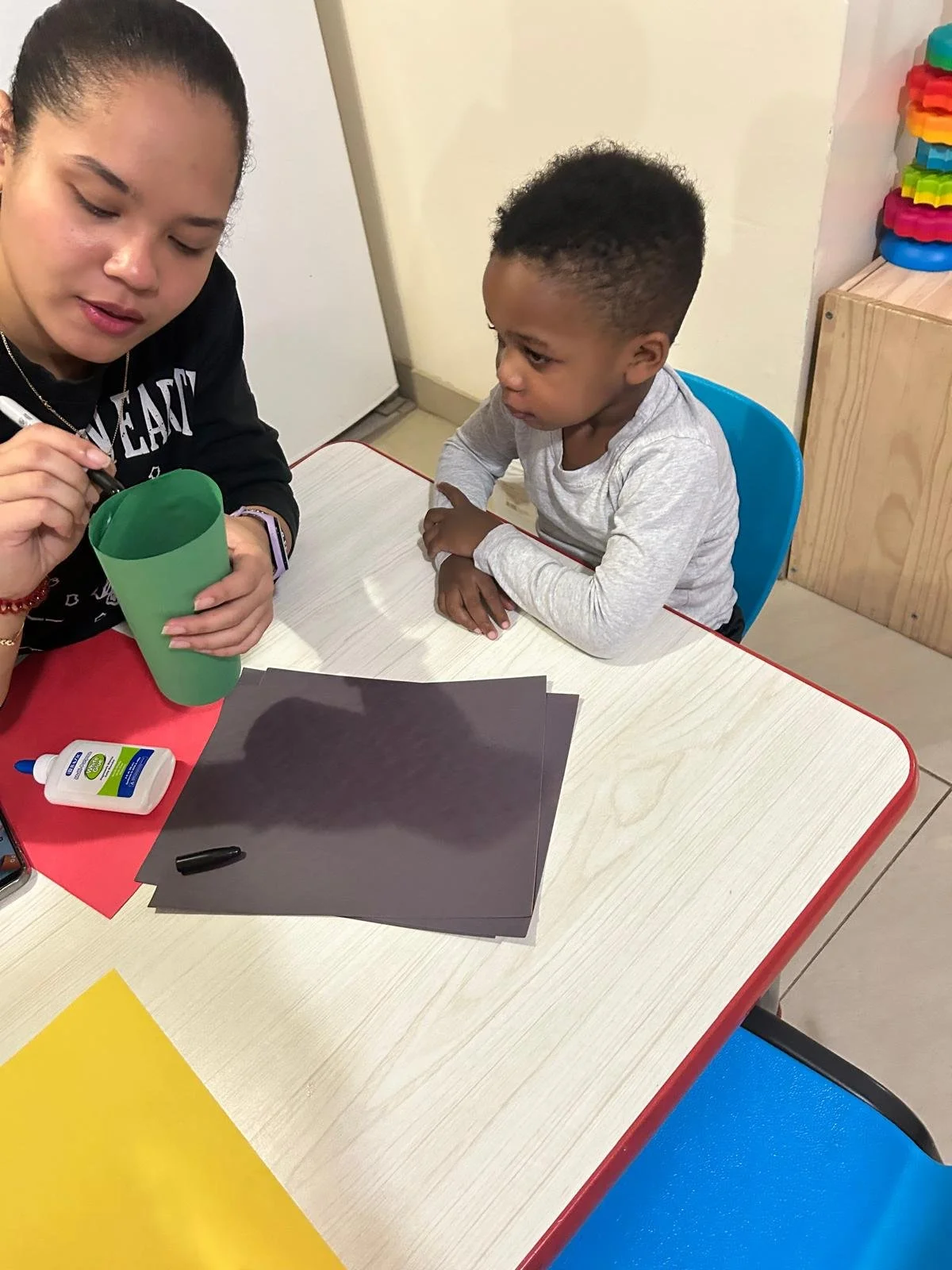 A caretaker is helping a young boy with a craft project at a table, with supplies like paper, glue, and a green cup.