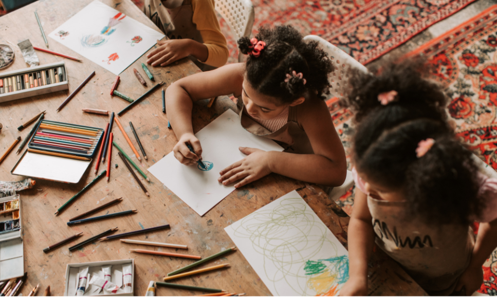 Three girls drawing and coloring at a table with scattered colored pencils and drawings in progress.
