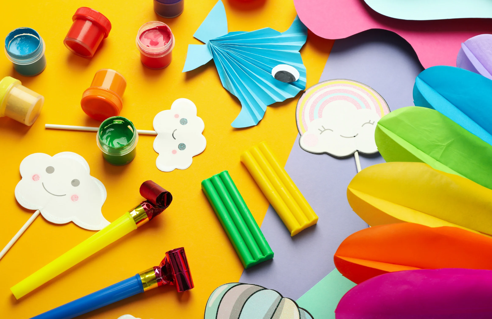 Colorful crafting supplies on a yellow background, including paint pots, paper cutouts of clouds and a rainbow, paper flowers, curling ribbons, and folded paper in various colors.