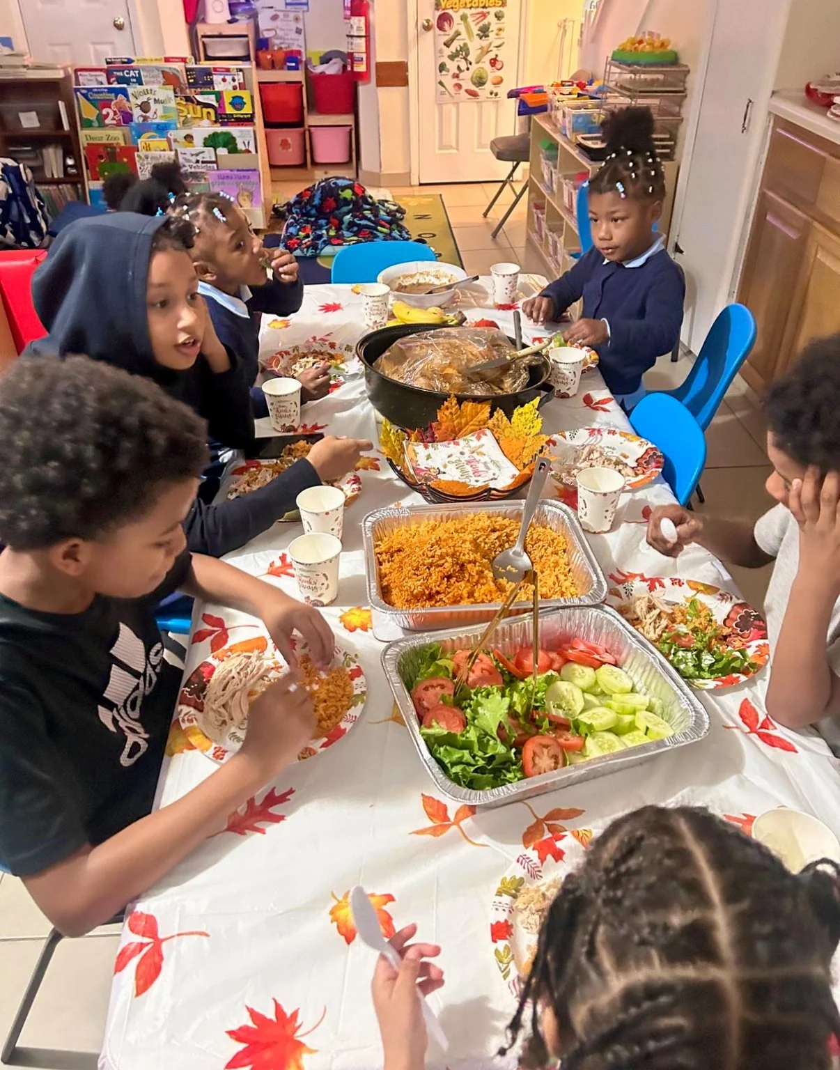 Children gathered around a table with Thanksgiving-themed tablecloth, enjoying a meal with rice, salad, and turkey in a classroom decorated with educational posters and books.