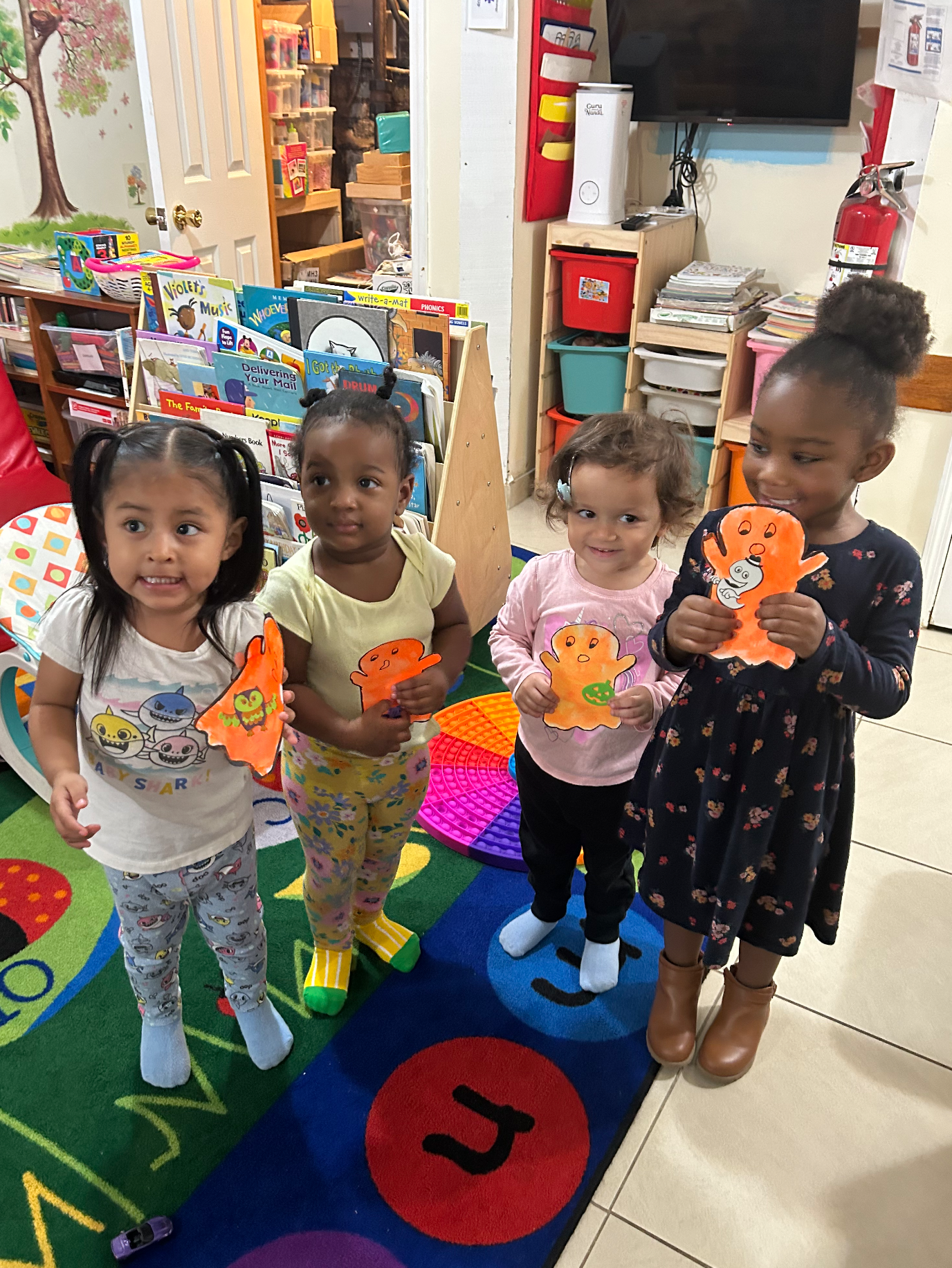 Four young girls standing together in a classroom, holding Halloween-themed paper crafts, smiling and posing for the photo.