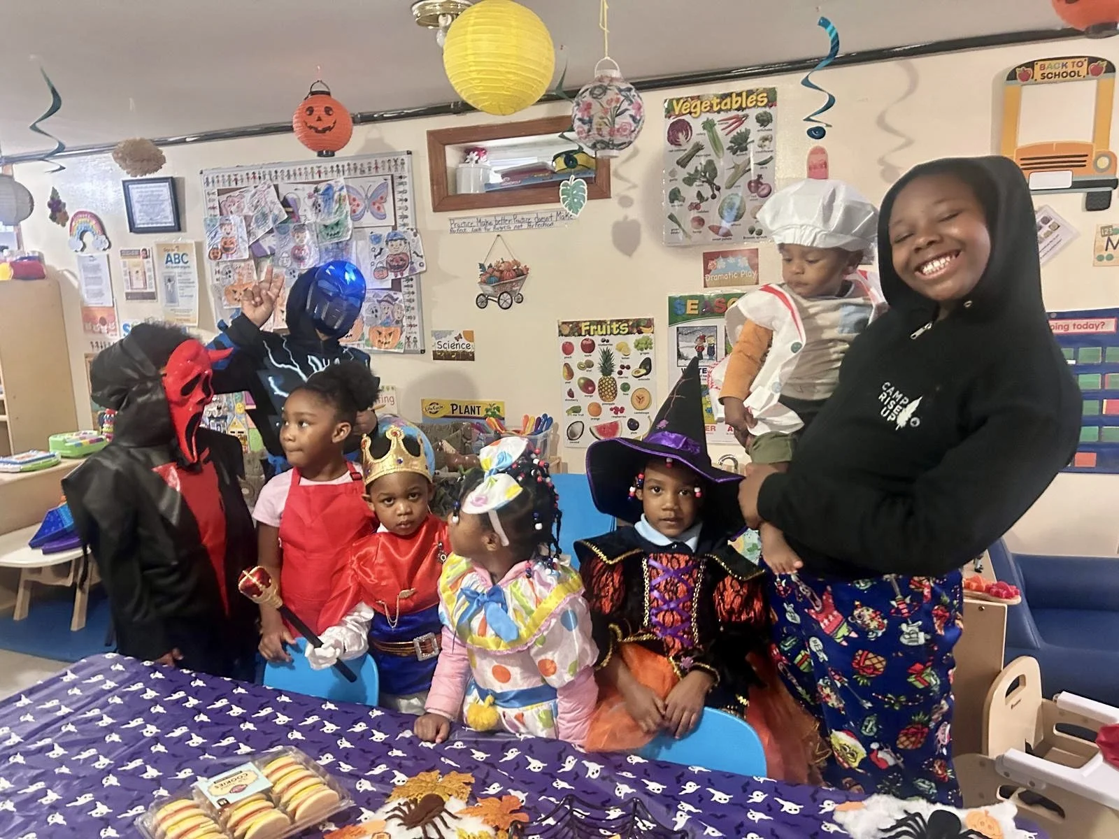 Children dressed in Halloween costumes at a birthday party, with a superhero, princess, witch, and other costumes, in a decorated room with Halloween and back-to-school decorations.