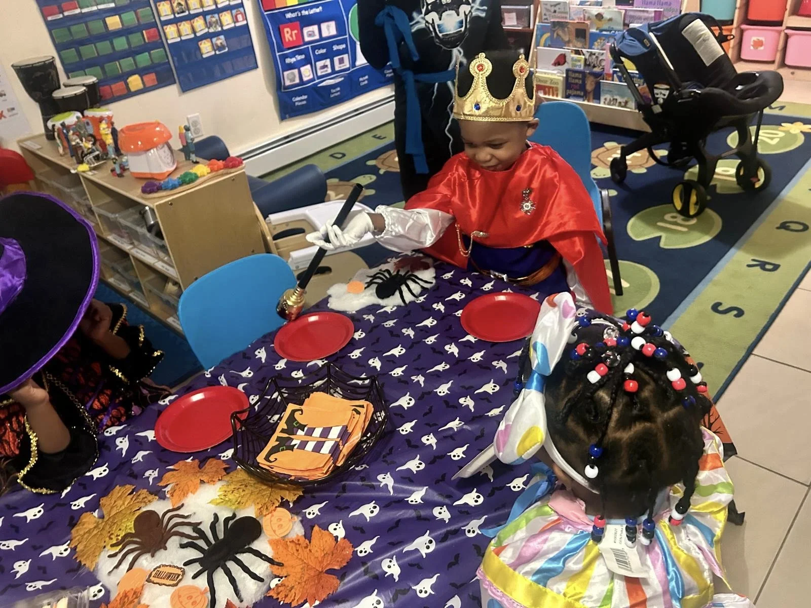 Two young children dressed in Halloween costumes sit at a decorated table in a classroom. One child is dressed as a king with a crown and red cape, holding a stick, while the other is wearing a colorful outfit with braided hair and beads. The table is adorned with orange leaves, toy spiders, and a small basket of Halloween-themed napkins. The classroom background features educational posters, toys, and storage bins.