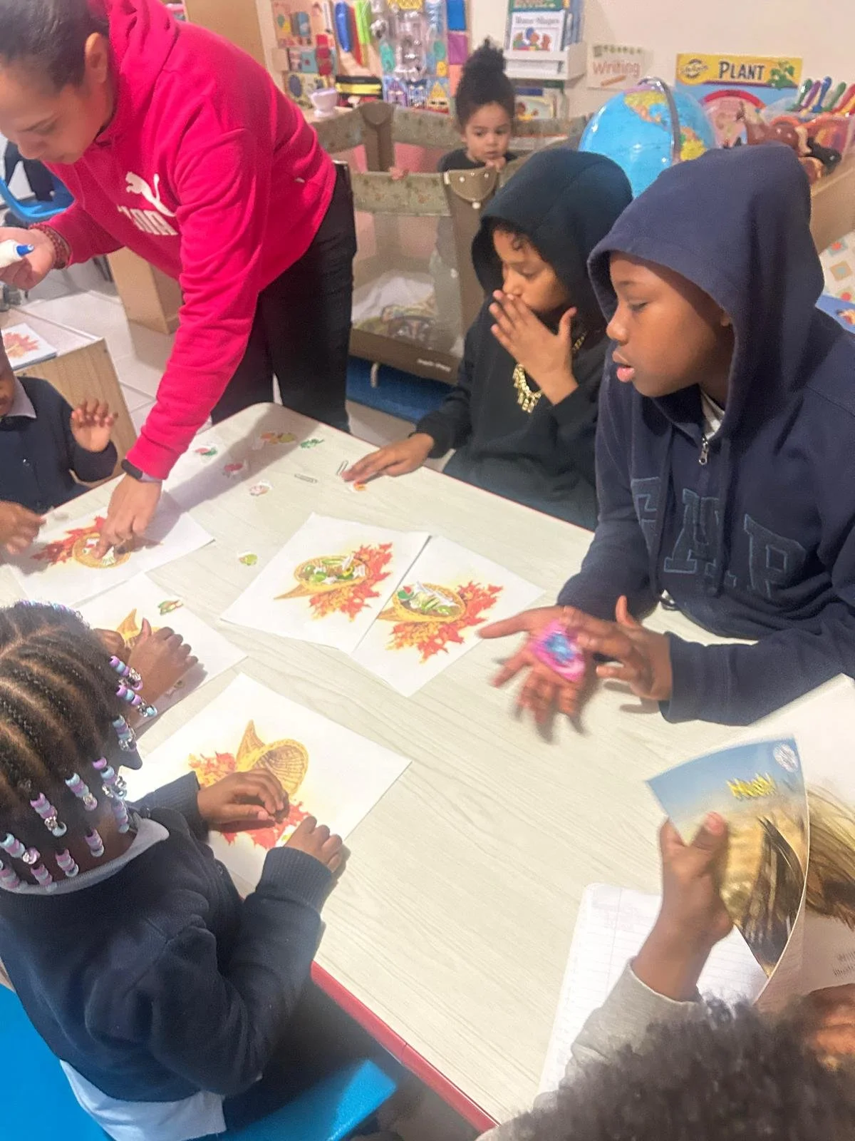 Children and a teacher working on Halloween-themed crafts at a table, with Halloween images on papers and various classroom decorations in the background.