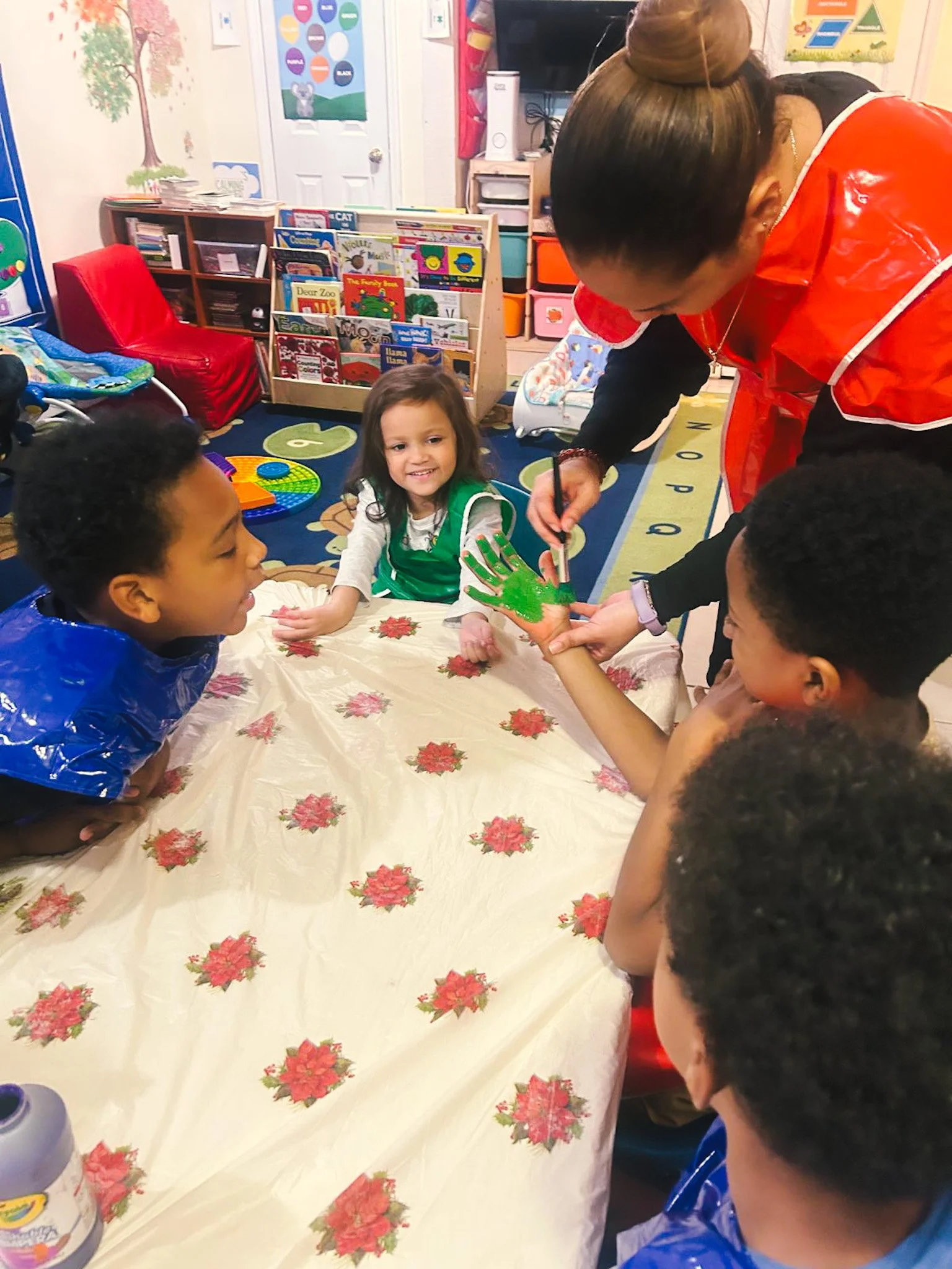 A group of young children sitting around a decorated table in a classroom, with a woman helping a girl paint a hand with green paint for a craft activity.