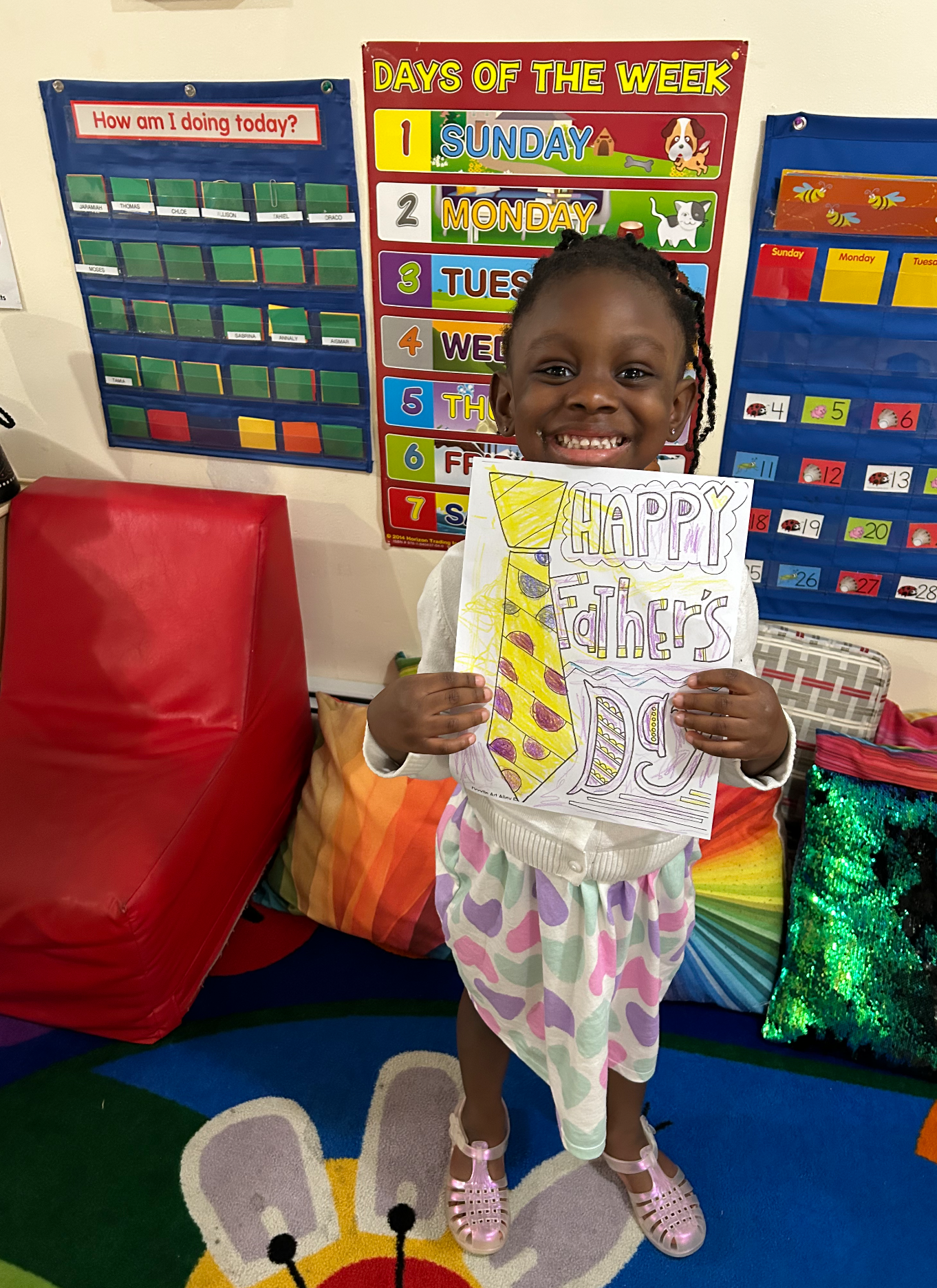 A young girl is smiling and holding a colorful Father's Day card inside a classroom. The card has a drawing of a tie and the words "Happy Father's" on it. Behind her, there are educational posters on the wall, including a days of the week chart and a behavior chart. The floor has a cheerful, animal-themed carpet.