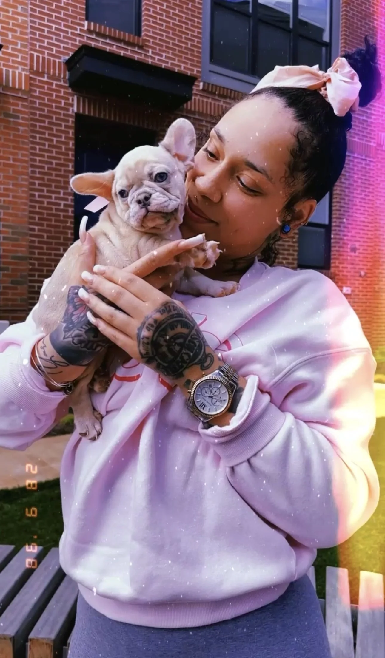 A woman holding a French Bulldog puppy outside in front of a brick building, smiling and looking at the puppy.