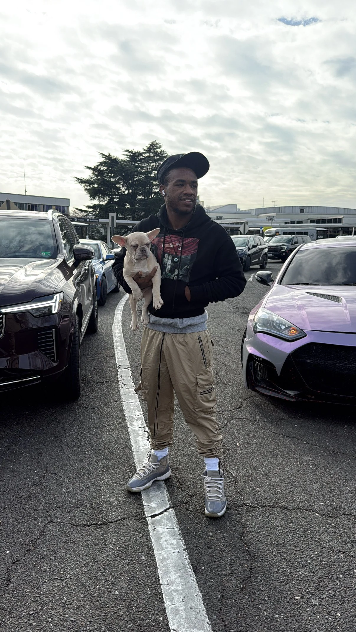 Young man in beige joggers and sneakers holding a small bulldog in a parking lot with luxury cars around, under a cloudy sky.