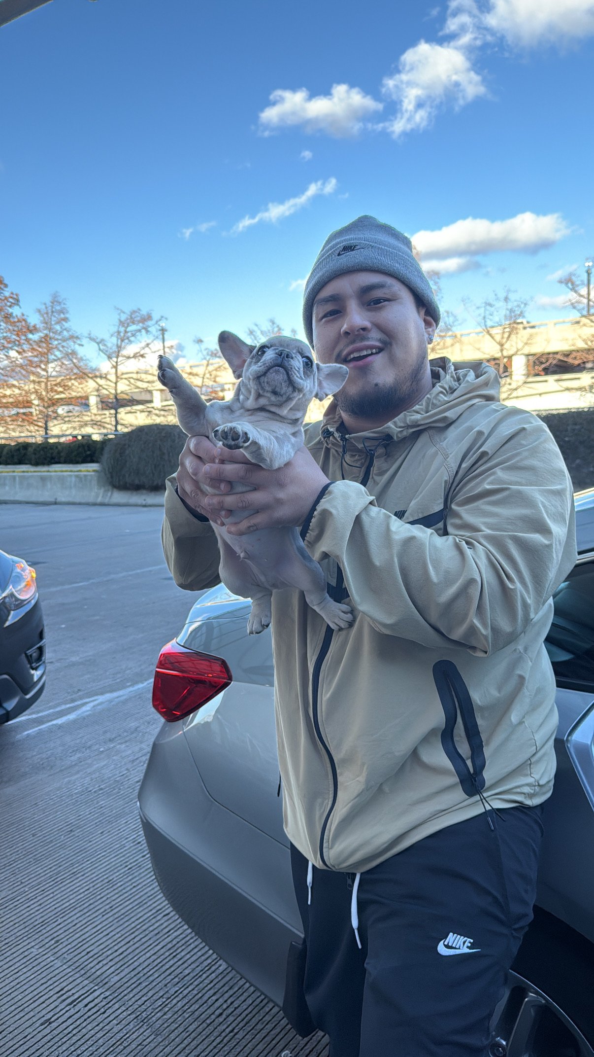 A man wearing a gray beanie and a beige jacket holding a small French bulldog puppy outside in a parking lot with a blue sky and a few clouds.