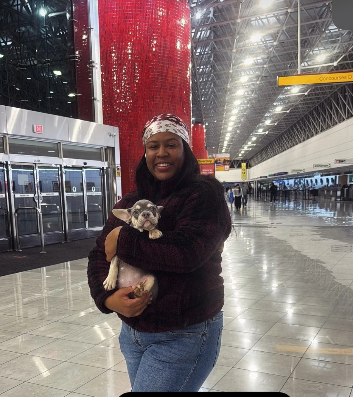 A woman smiling while holding a small puppy inside an airport terminal with high ceilings, check-in counters, and yellow signs in the background.