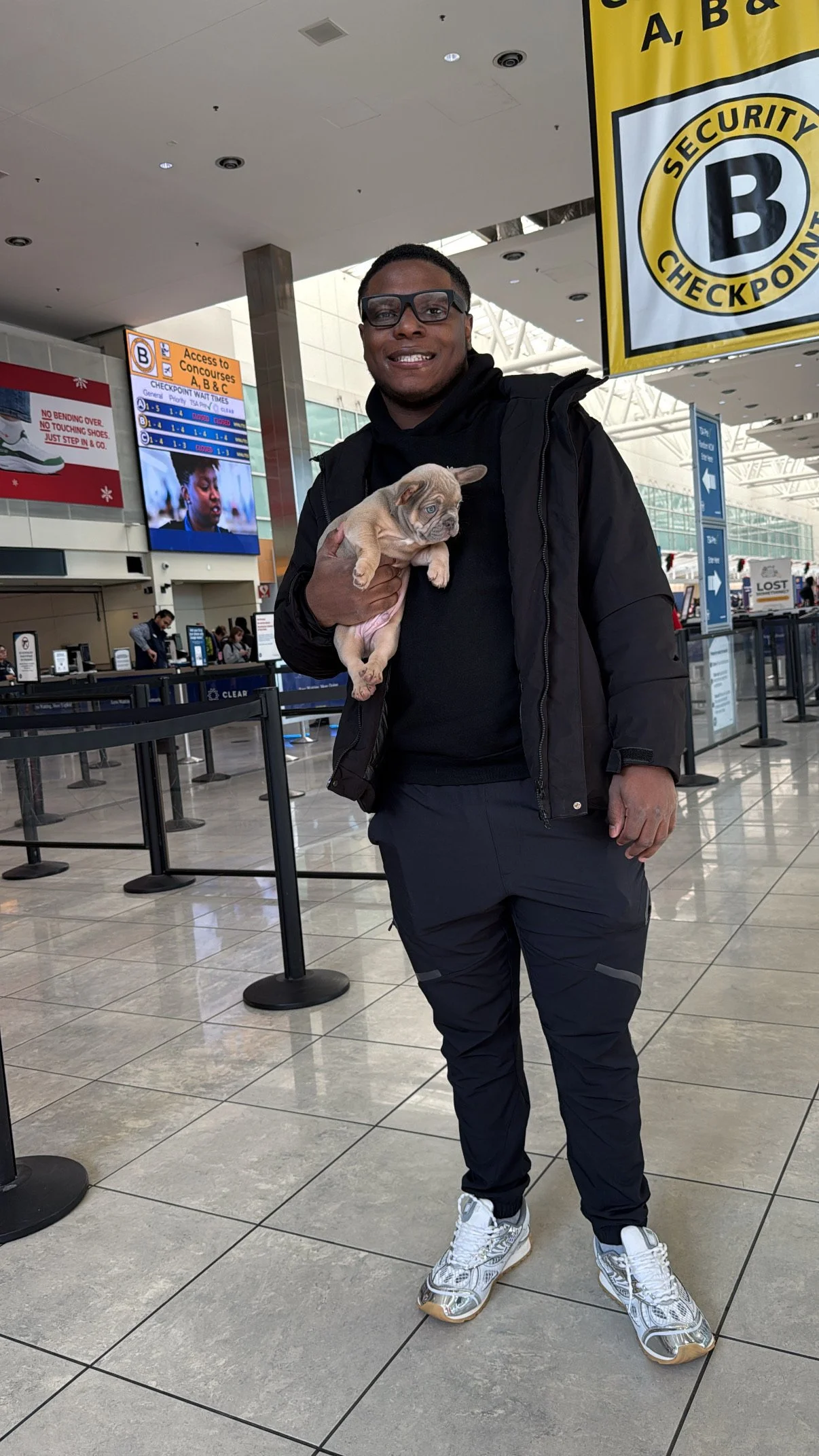 A man wearing glasses, a black jacket, and black pants is holding a small puppy in his arm at an airport terminal. There are signs and counters in the background.